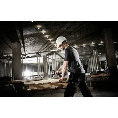Ein Bauarbeiter mit Schutzhelm sägt Holz auf einer Baustelle. Der Hintergrund zeigt unvollständige Betonstrukturen und schwaches Licht.