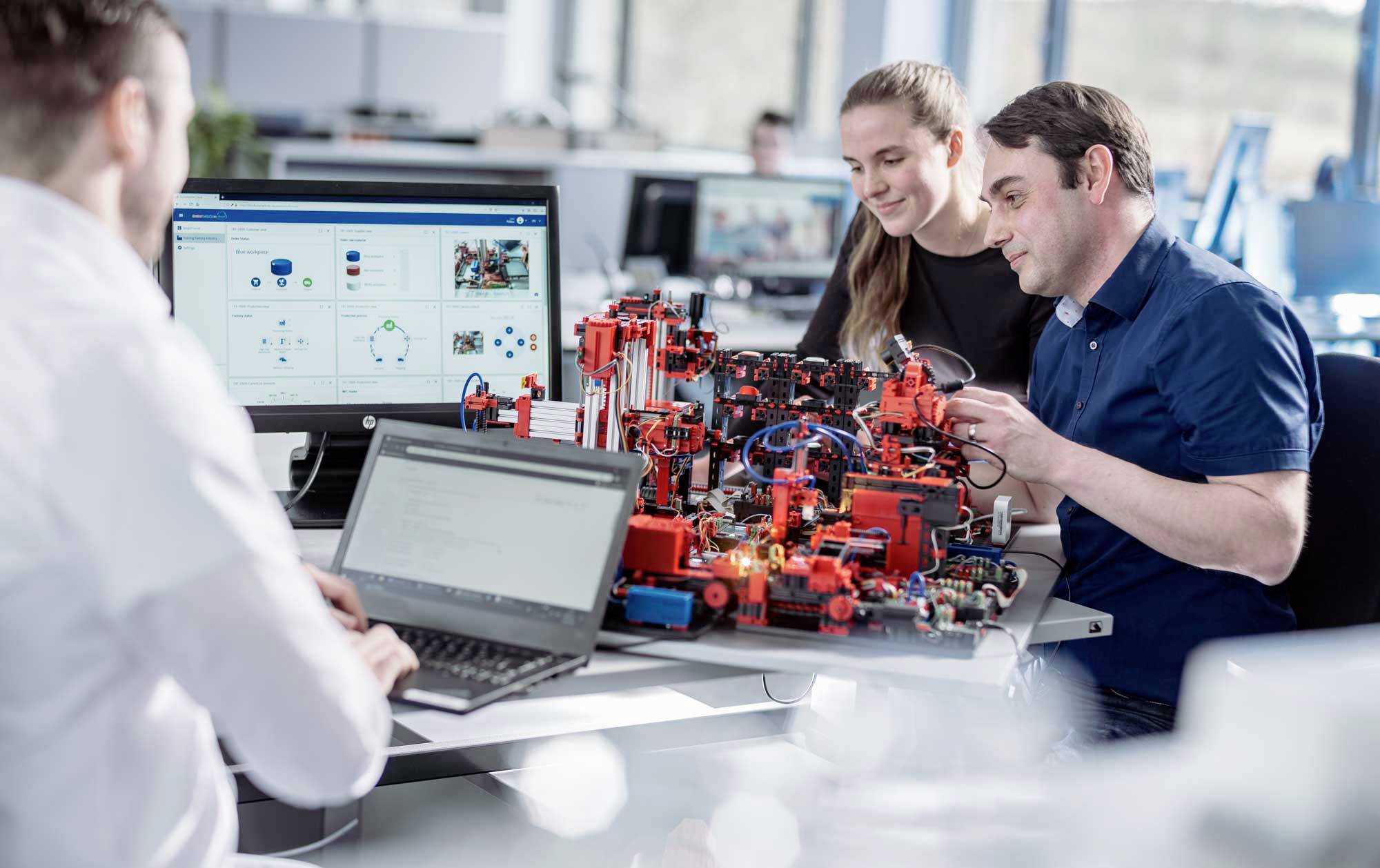 Drei Personen arbeiten gemeinsam an einem Tisch an einem technischen Projekt mit Laptops. Im Hintergrund sind Bildschirme sichtbar.