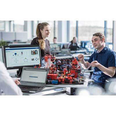 Drei Personen diskutieren über ein technisches Modell auf einem Tisch in einem Büro. Auf den Monitoren sind Diagramme und Daten zu sehen.