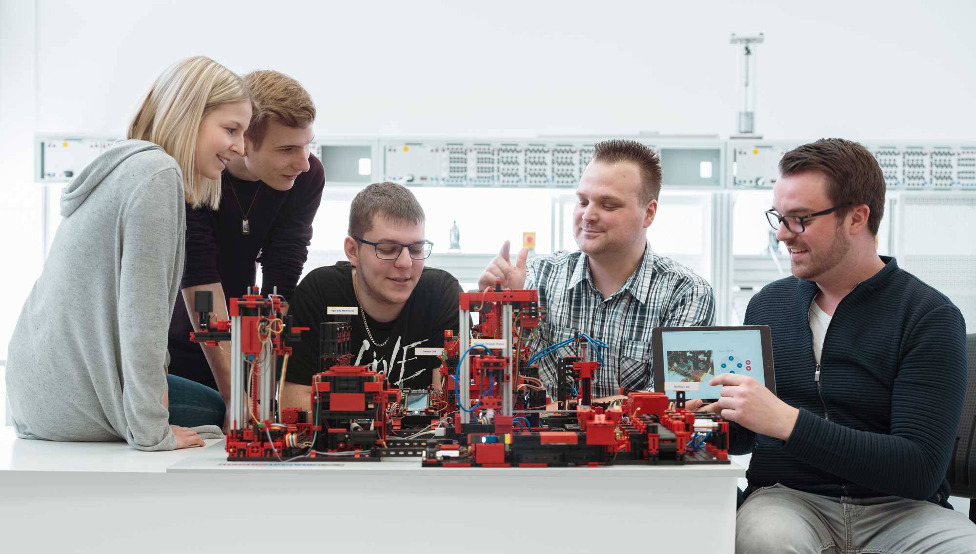 Eine Gruppe von fünf Personen arbeitet gemeinsam an einem technischen Modell aus roten und schwarzen Teilen auf einem weißen Tisch.