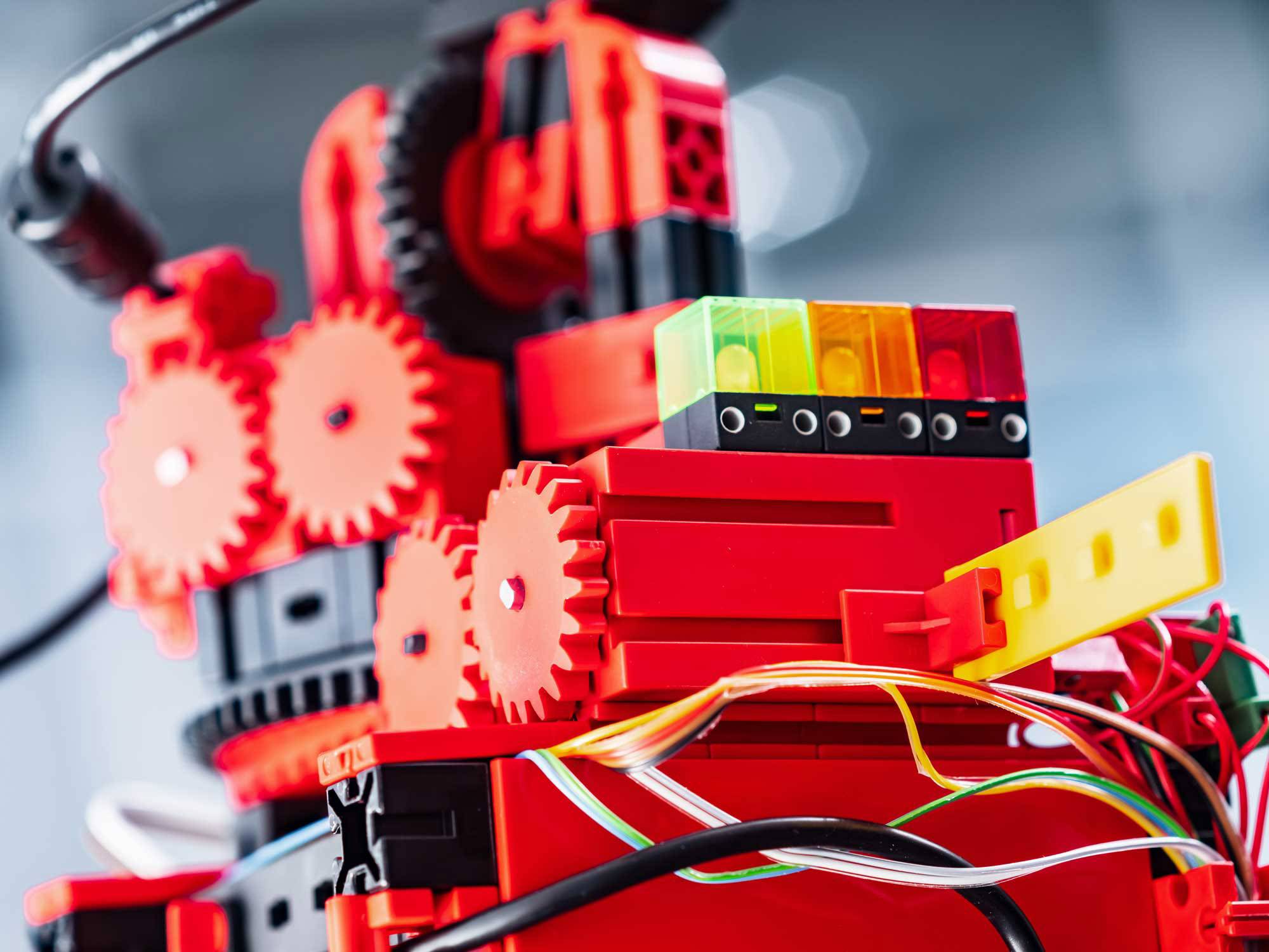 Ein leuchtend roter Roboter mit Zahnrädern und Kabeln. Bunte Steckmodule oben; Technik und Kreativität symbolisierend.