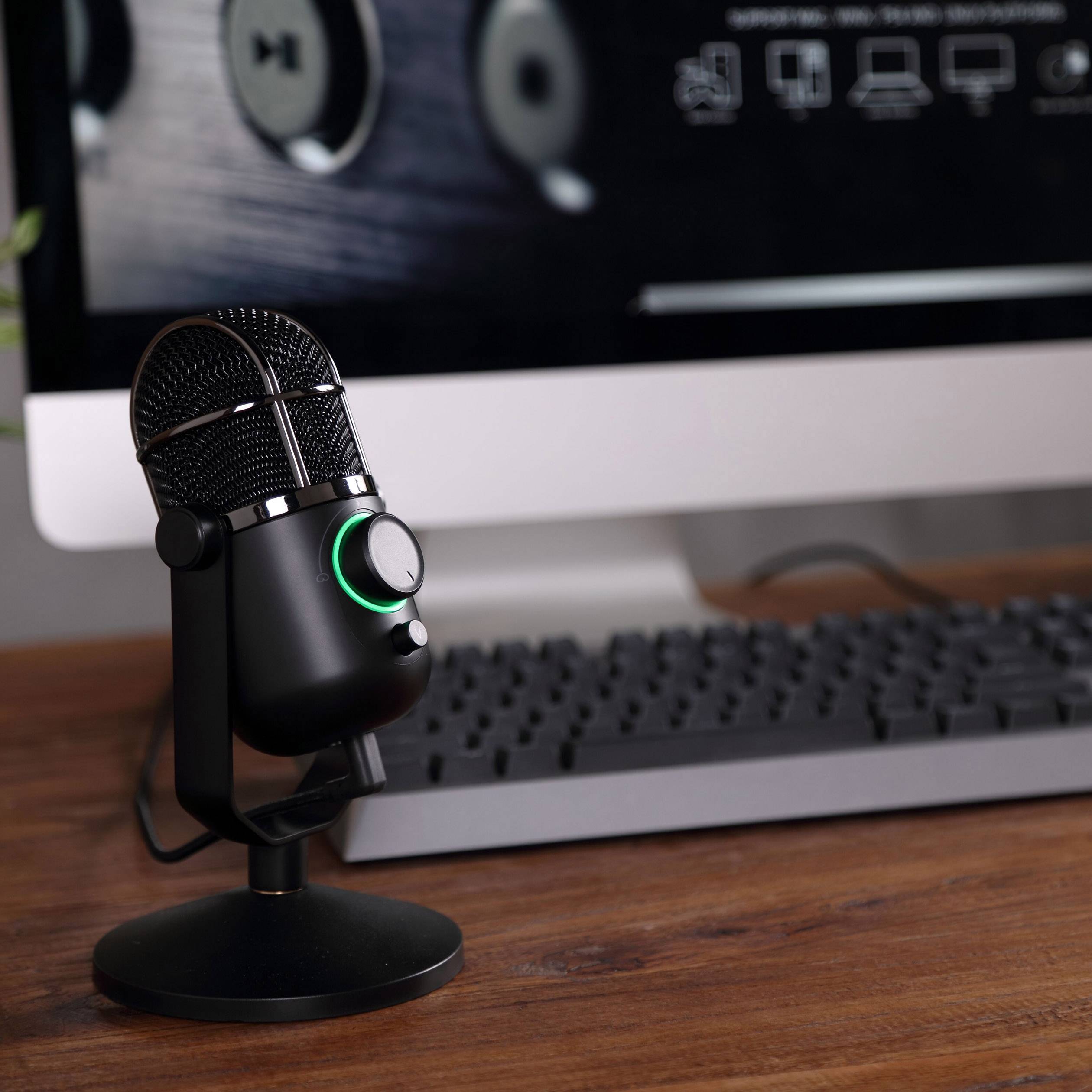 Ein Mikrofon steht auf einem Schreibtisch vor einem Computerbildschirm und einer Tastatur. Das Mikrofon ist eingeschaltet.