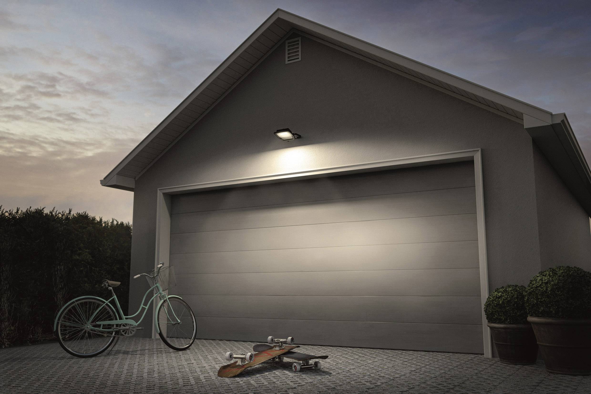 Ein weißes Fahrrad und ein Skateboard liegen vor einer geschlossenen Garagentür in der Dämmerung. Eine Lampe beleuchtet die Szene.