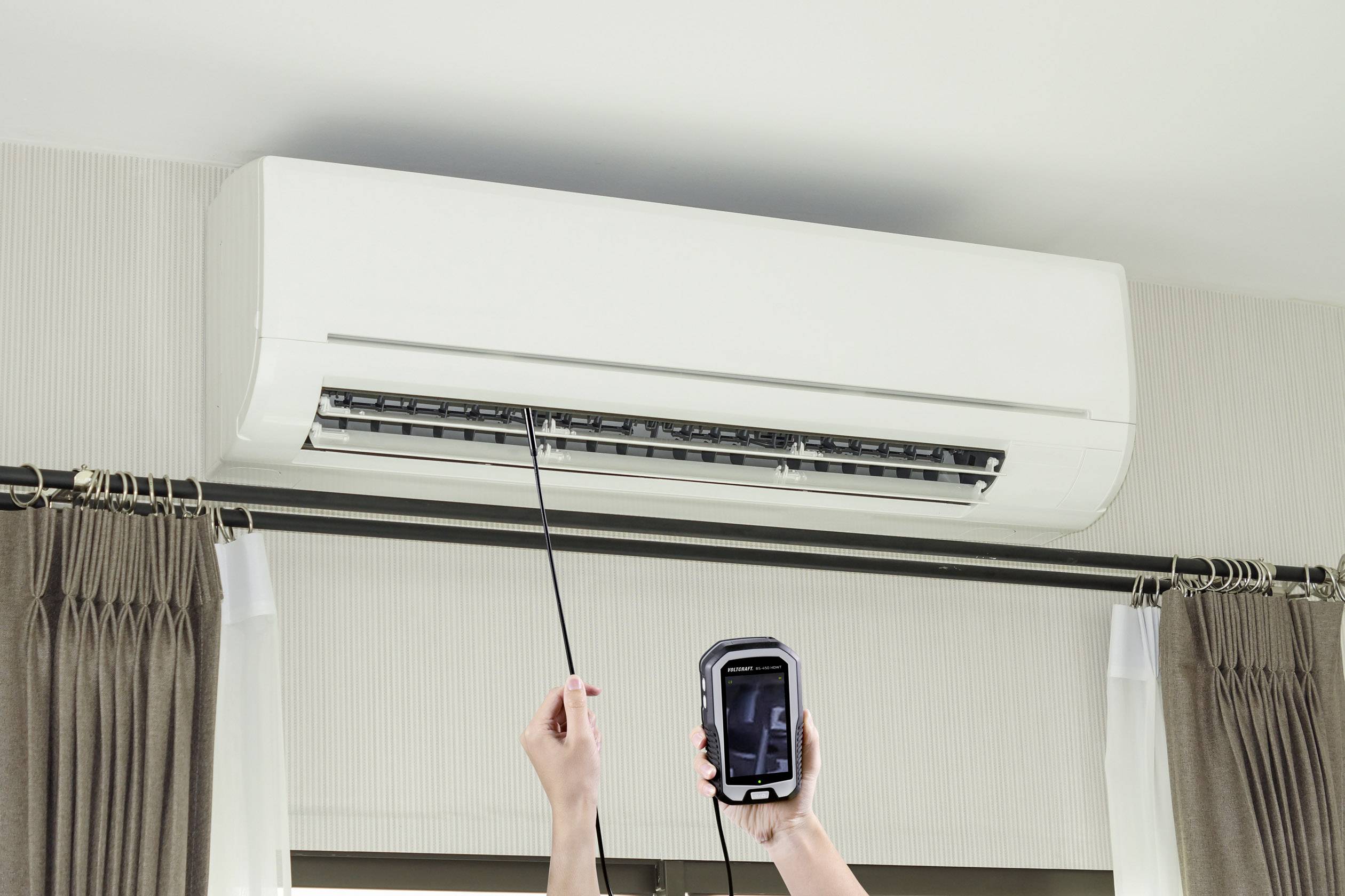 Eine Person prüft mit einem Gerät die Funktion einer Klimaanlage an der Wand. Die Hände der Person sind sichtbar, während sie das Gerät hält.