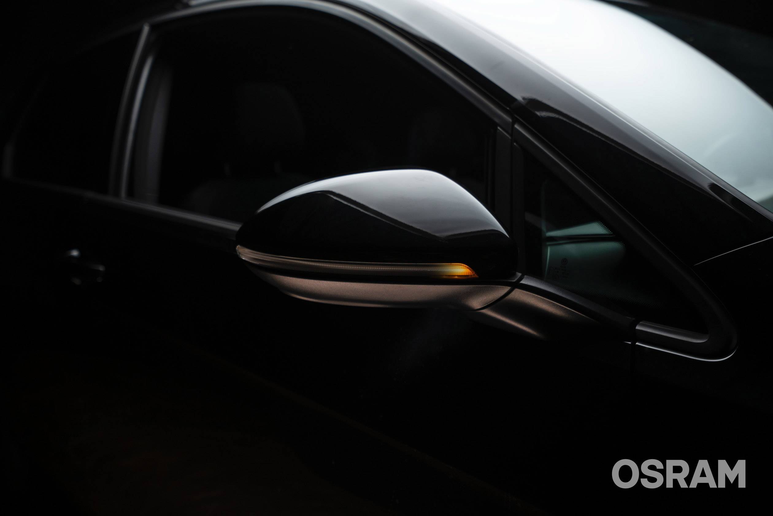 Ein Seitenspiegel eines Autos mit eingeschaltetem Blinker, in einem dunklen Ambiente. Markenname 'OSRAM' unten rechts sichtbar.