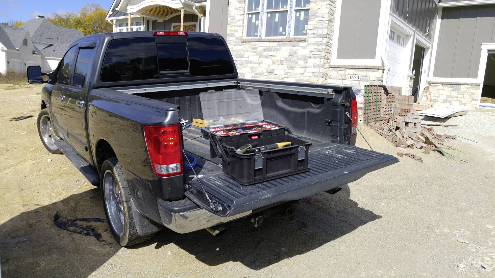 Schwarzer Pickup-Truck mit geöffneter Ladefläche, auf der Werkzeuge und eine schwarze Box stehen, vor einem im Bau befindlichen Haus.