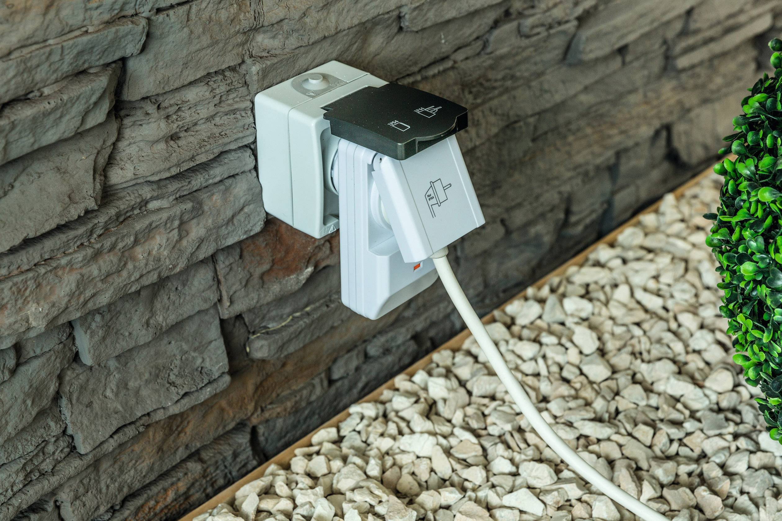 Eine weiße Steckdose mit einem eingesteckten Stecker befindet sich an einer Steinwand. Ein Kabel führt zu einem Elektrogerät.