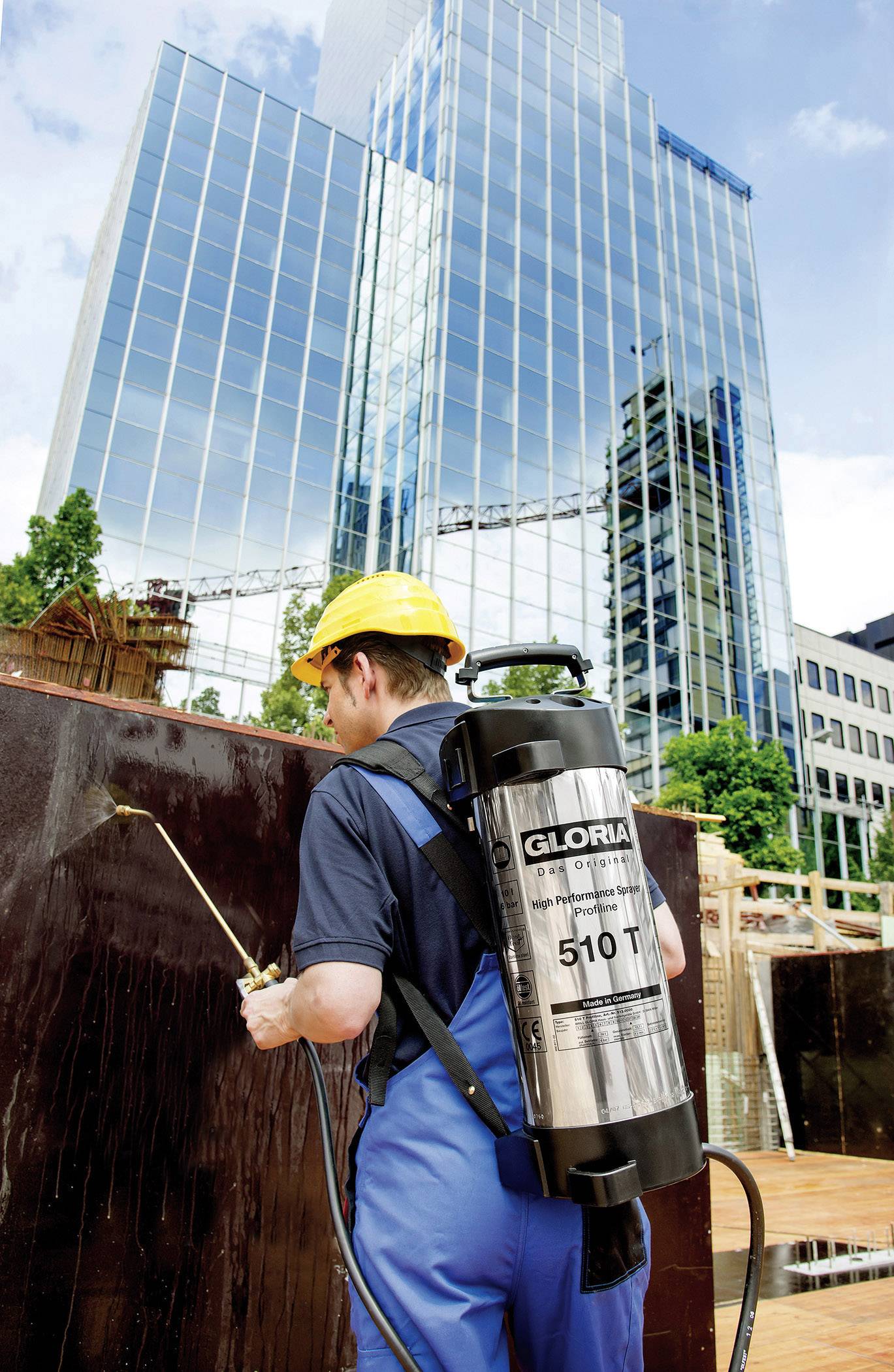Person in blauer Arbeitskleidung und gelbem Helm besprüht mit einem Tank auf dem Rücken eine Wand auf einer Baustelle vor einem Hochhaus.