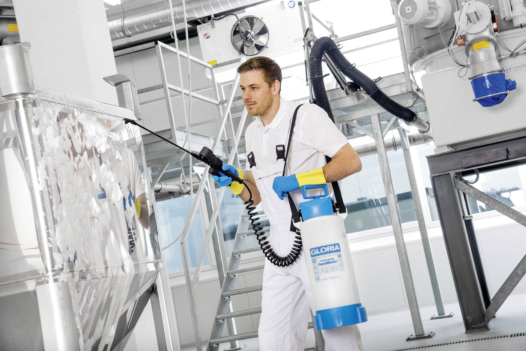 Ein Mann in Arbeitskleidung reinigt mit einem Hochdruckreiniger eine metallene Industrieanlage in einer Fabrikumgebung.
