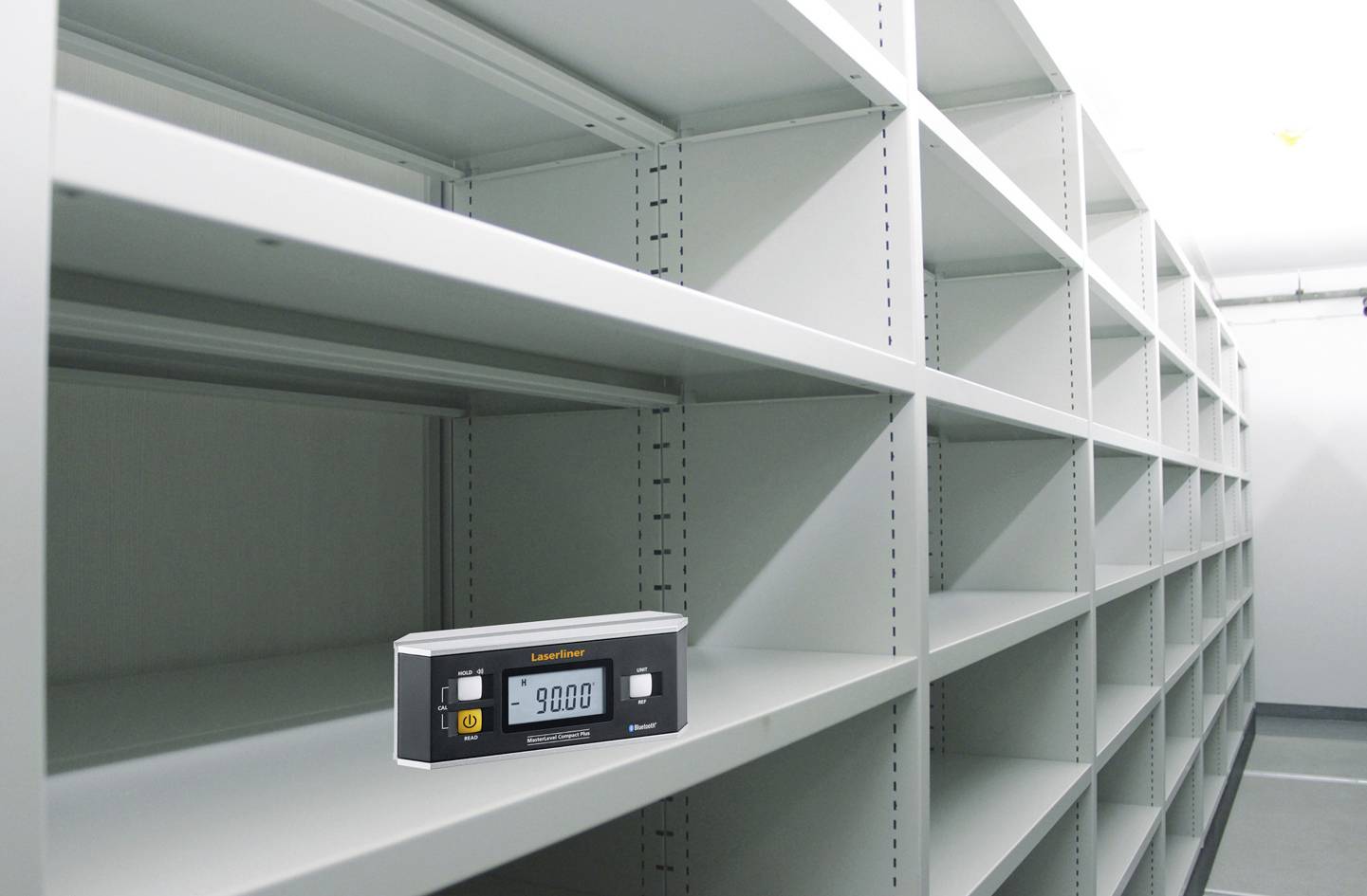 Messgerät mit digitalem Display zeigt '9.000'. Es liegt vor leeren, grauen Regalen in einem Lagerraum.