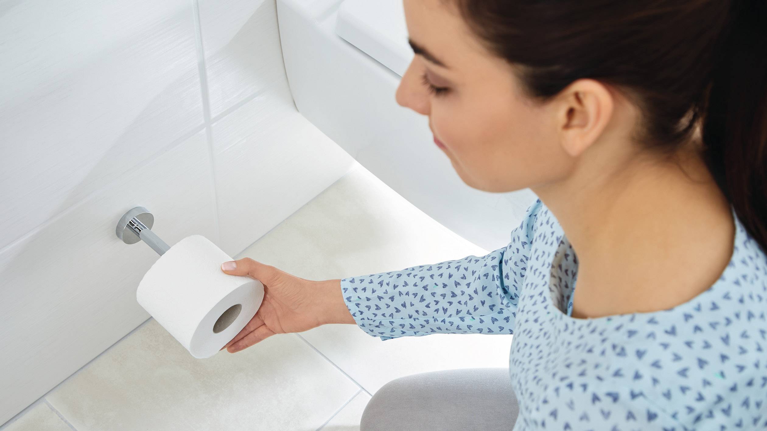 Eine Person sitzt in einem Badezimmer und hält eine Toilettenpapierrolle in der Hand, bereit sie auf den Halter zu stecken.
