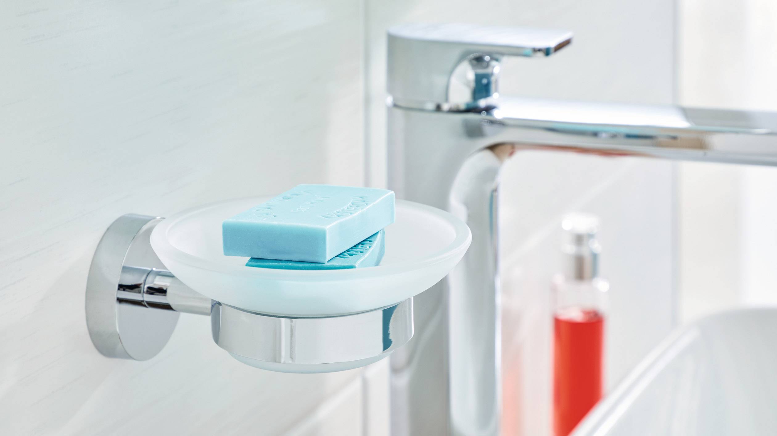 Blaue Seife liegt in einer Seifenschale neben einem modernen Wasserhahn im Badezimmer. Im Hintergrund steht ein Behälter mit roter Flüssigkeit.