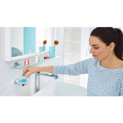 Eine Frau im Badezimmer nimmt ein Stück Seife von einer Halterung. Auf einem Regal stehen Kosmetikartikel und Bürsten.