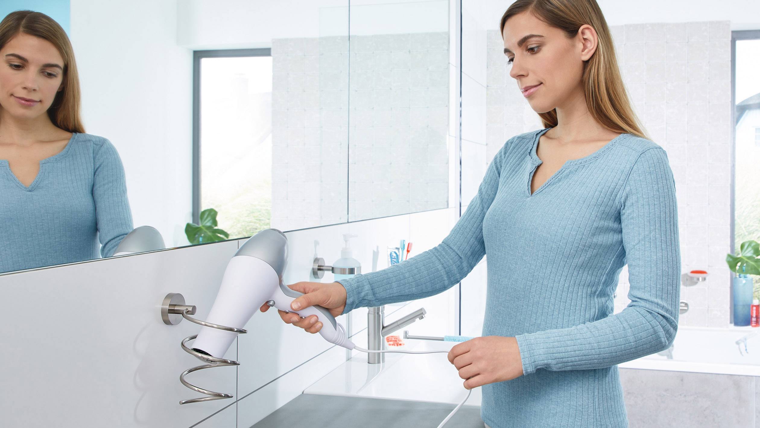 Eine Frau föhnt ihre Haare im Badezimmer. Sie trägt einen blauen Pullover und steht vor einem Spiegel. Im Hintergrund sind Badezimmerutensilien sichtbar.