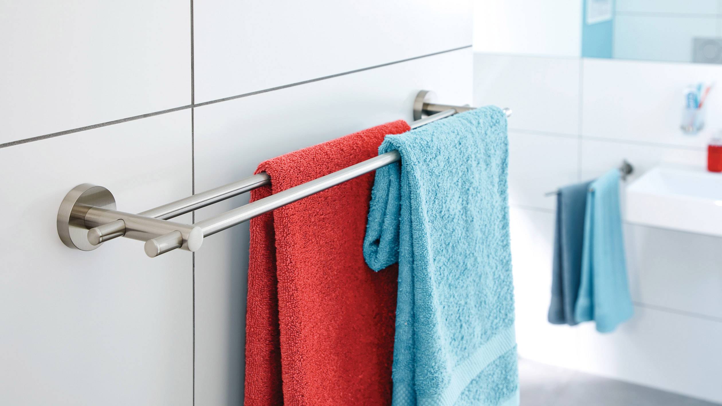 Zwei Handtücher, eines rot und eines blau, hängen auf einem Handtuchhalter im Badezimmer. Im Hintergrund ist ein Waschbecken zu sehen.