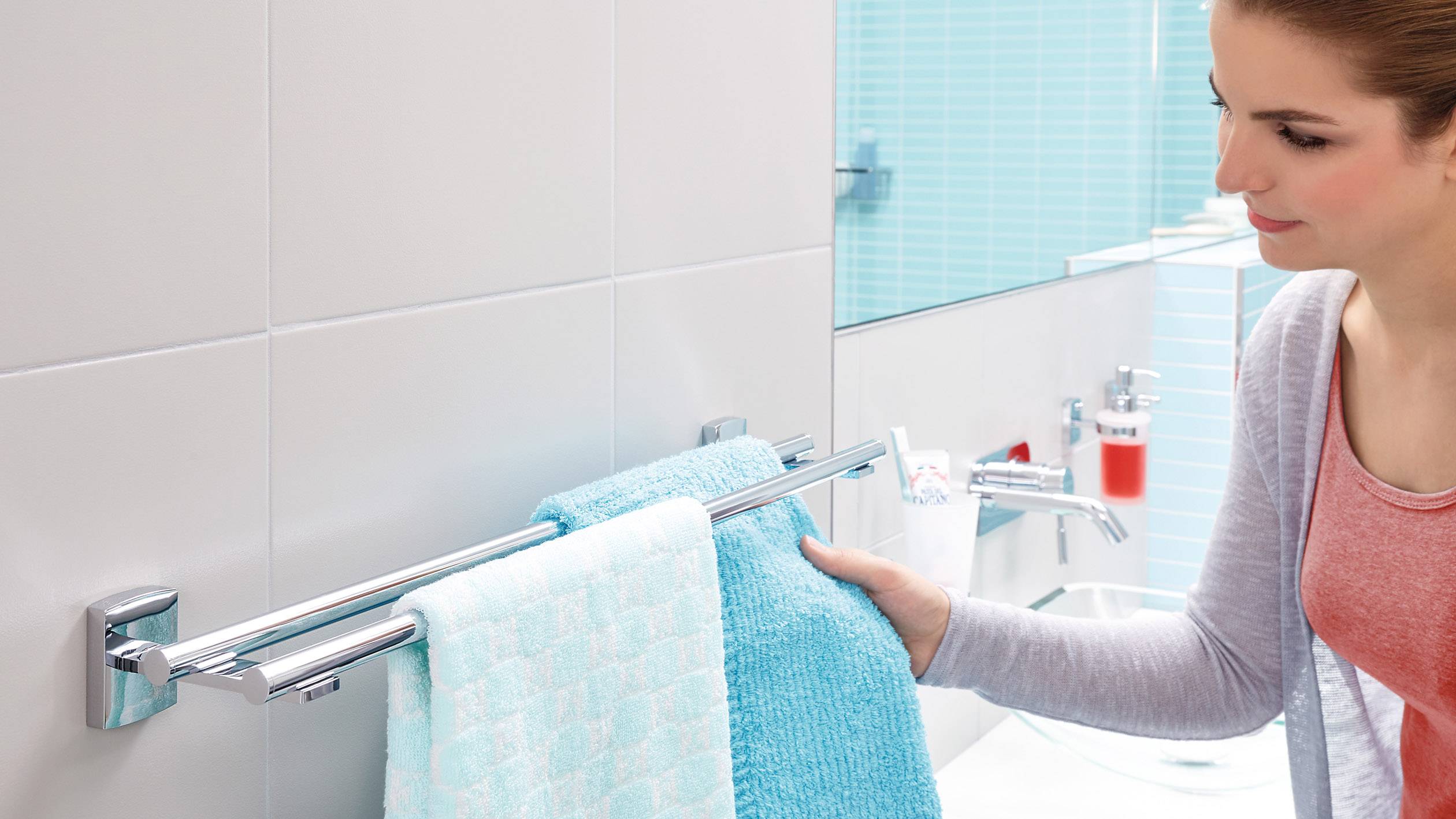 Eine Frau hängt ein blaues Handtuch neben einem hellgrünen Handtuch im Badezimmer auf. Im Hintergrund sind Fliesen und ein Waschbecken zu sehen.