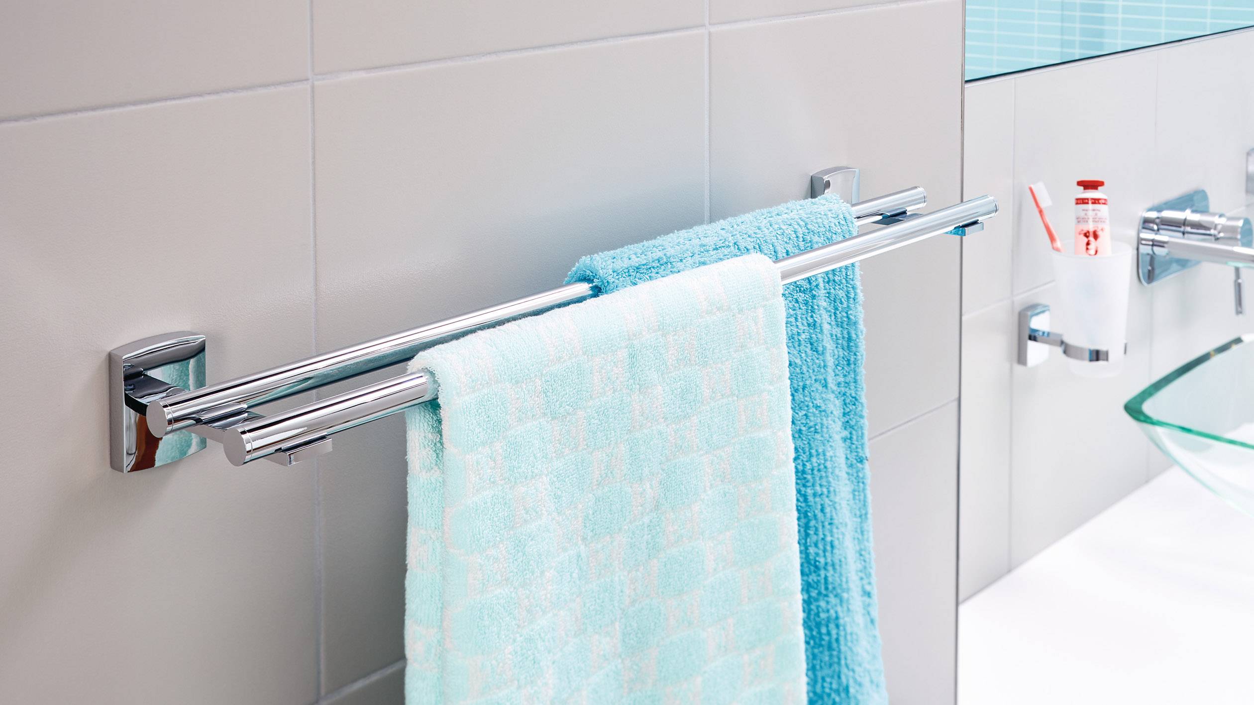 Ein Handtuchhalter mit zwei blauen Handtüchern hängt an einer Badezimmerwand. Im Hintergrund sieht man eine Tube Zahnpasta und ein Waschbecken.