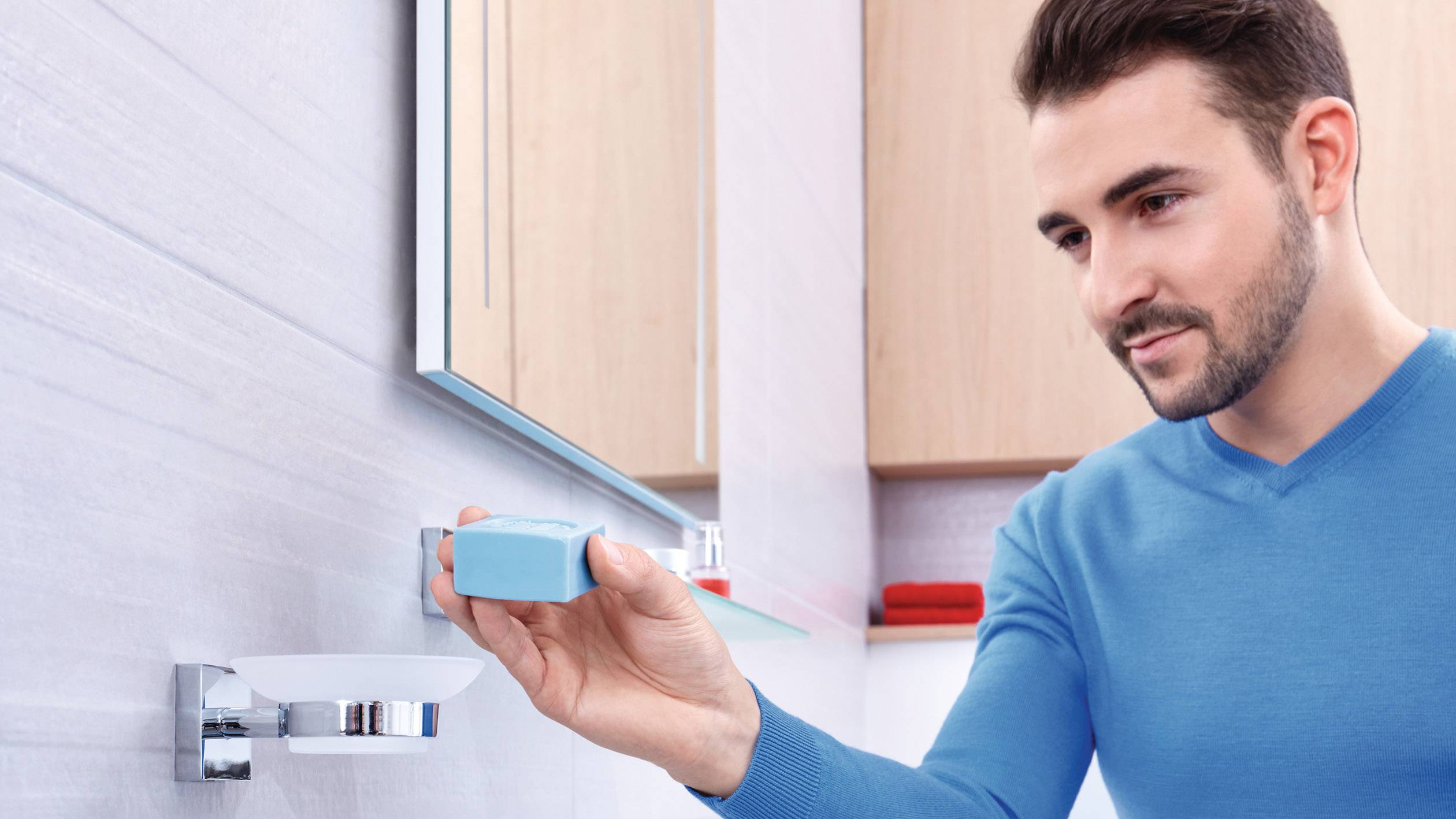 Ein Mann im blauen Pullover legt ein blaues Seifenstück auf einen Seifenhalter im Badezimmer.