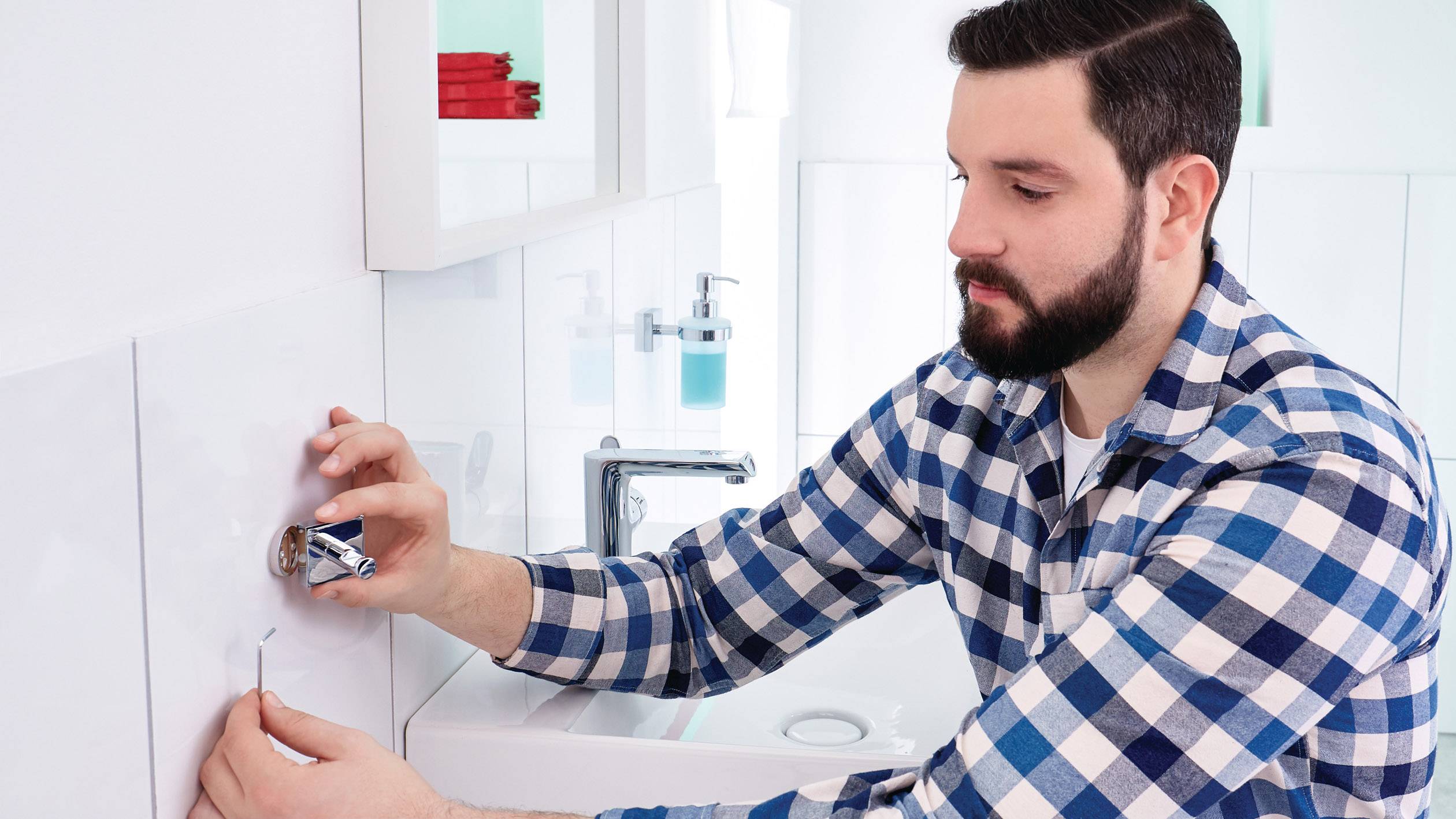 Ein Mann im karierten Hemd montiert eine Armatur im Badezimmer. Im Hintergrund sind ein Waschbecken und Pflegeprodukte sichtbar.