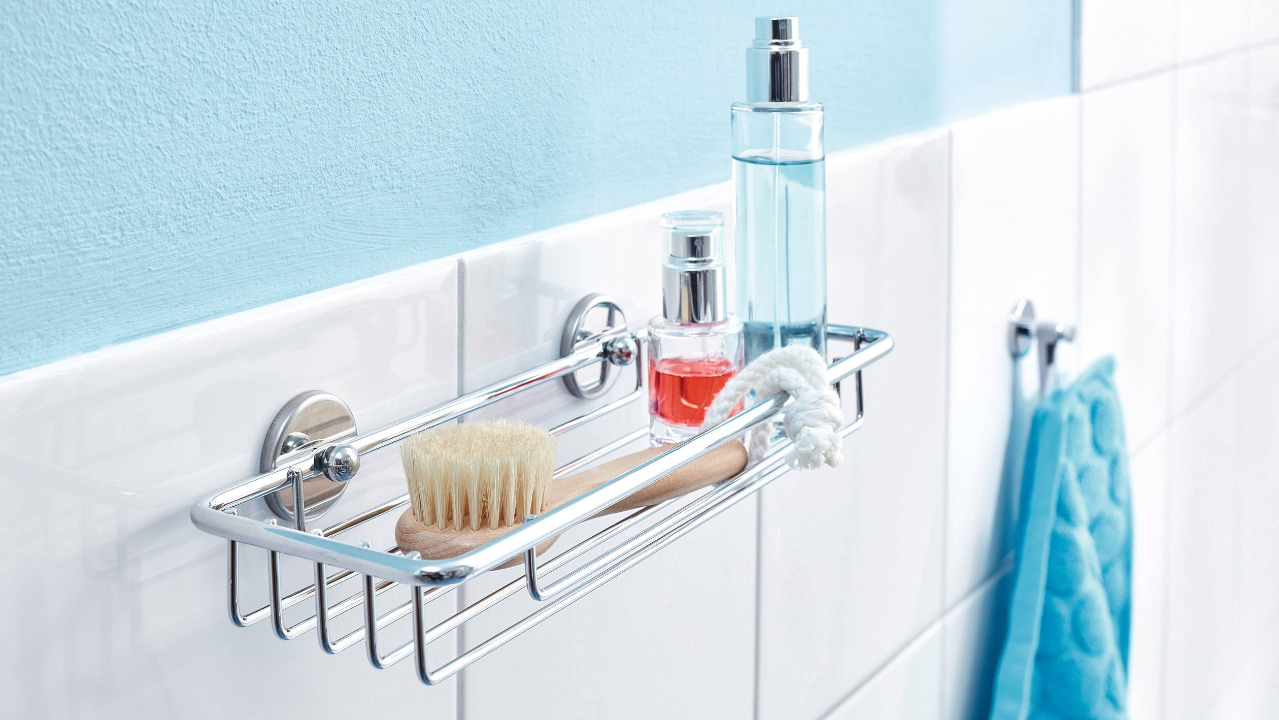 Ein Metallregal im Badezimmer mit Bürste, blauem und rotem Duschgel auf weißen Fliesen. Ein blaues Handtuch hängt daneben.