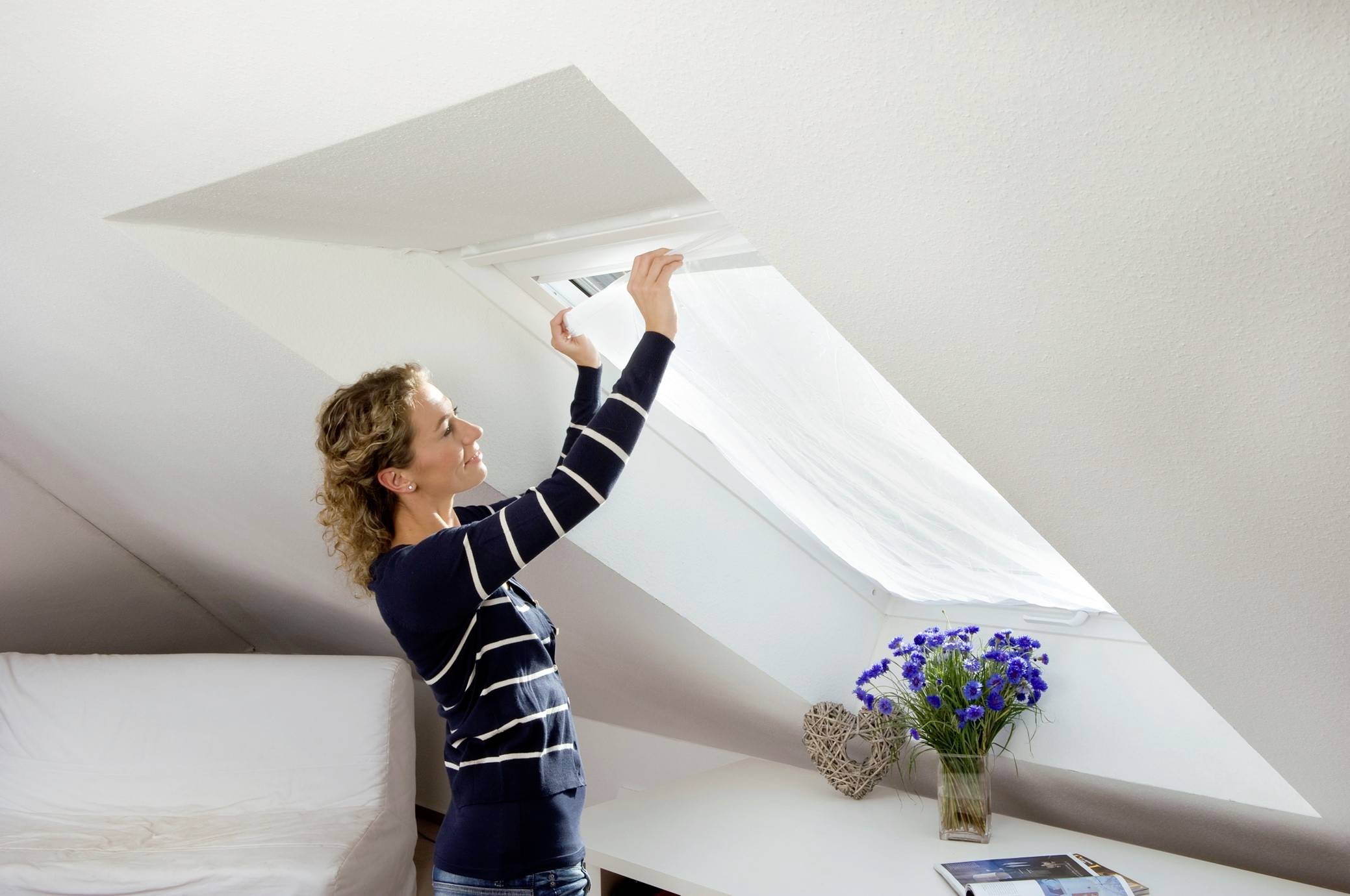 Eine Frau öffnet ein Dachfenster in einem hellen Zimmer. Auf einem Tisch darunter steht eine Vase mit blauen Blumen.