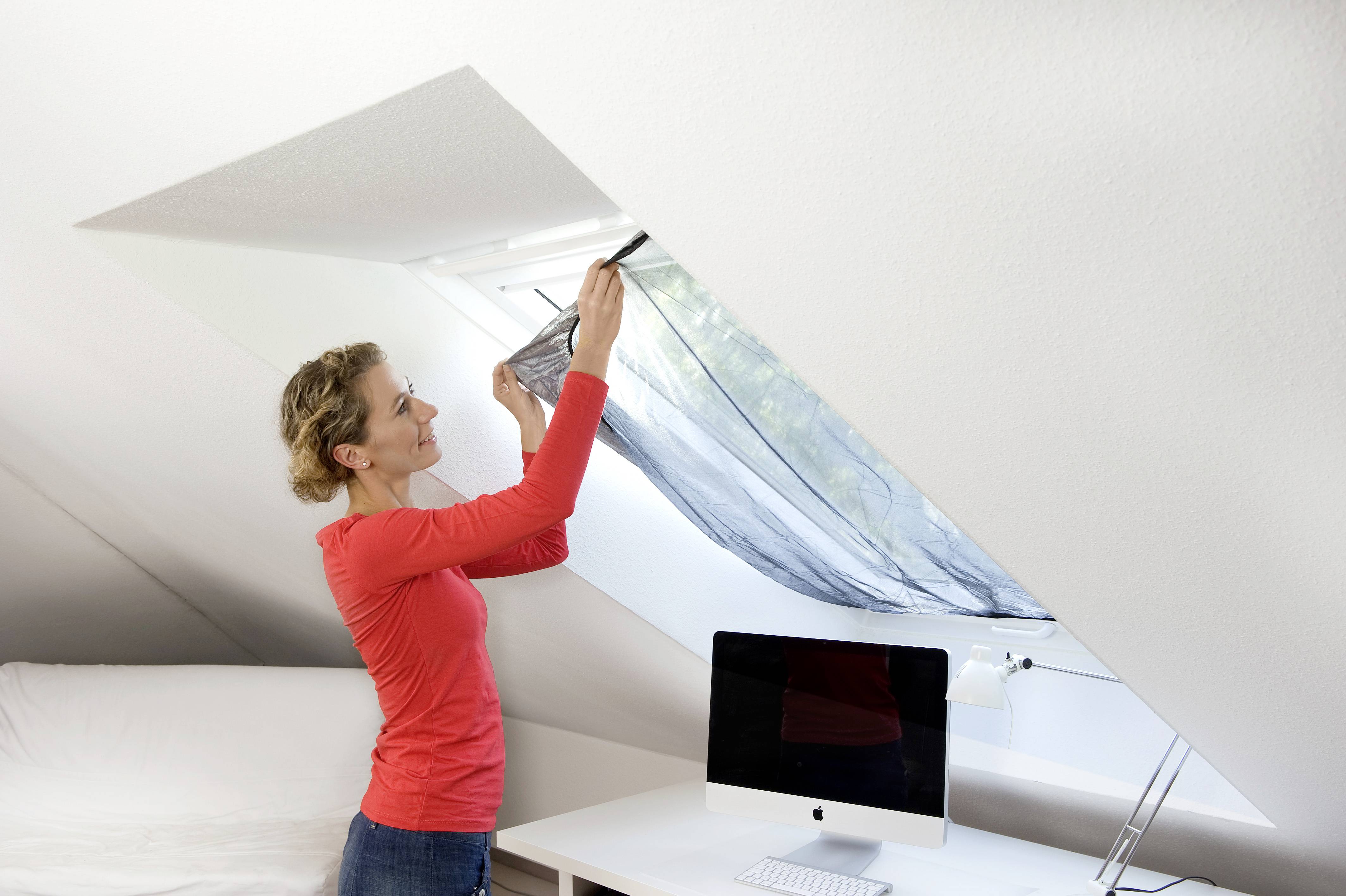 Eine Frau im roten Pullover zieht Vorhänge an einem Dachfenster im Büro mit einem Computer auf dem Schreibtisch zu.