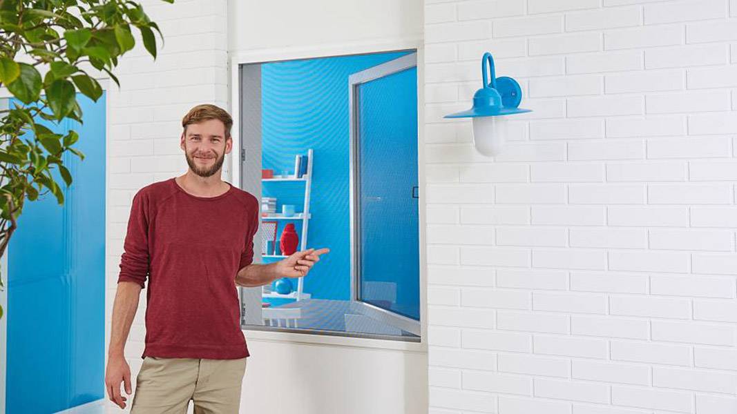 Ein lächelnder Mann in einem roten Pullover steht vor einer weißen Backsteinmauer mit einer blauen Lampe, zeigt nach rechts.