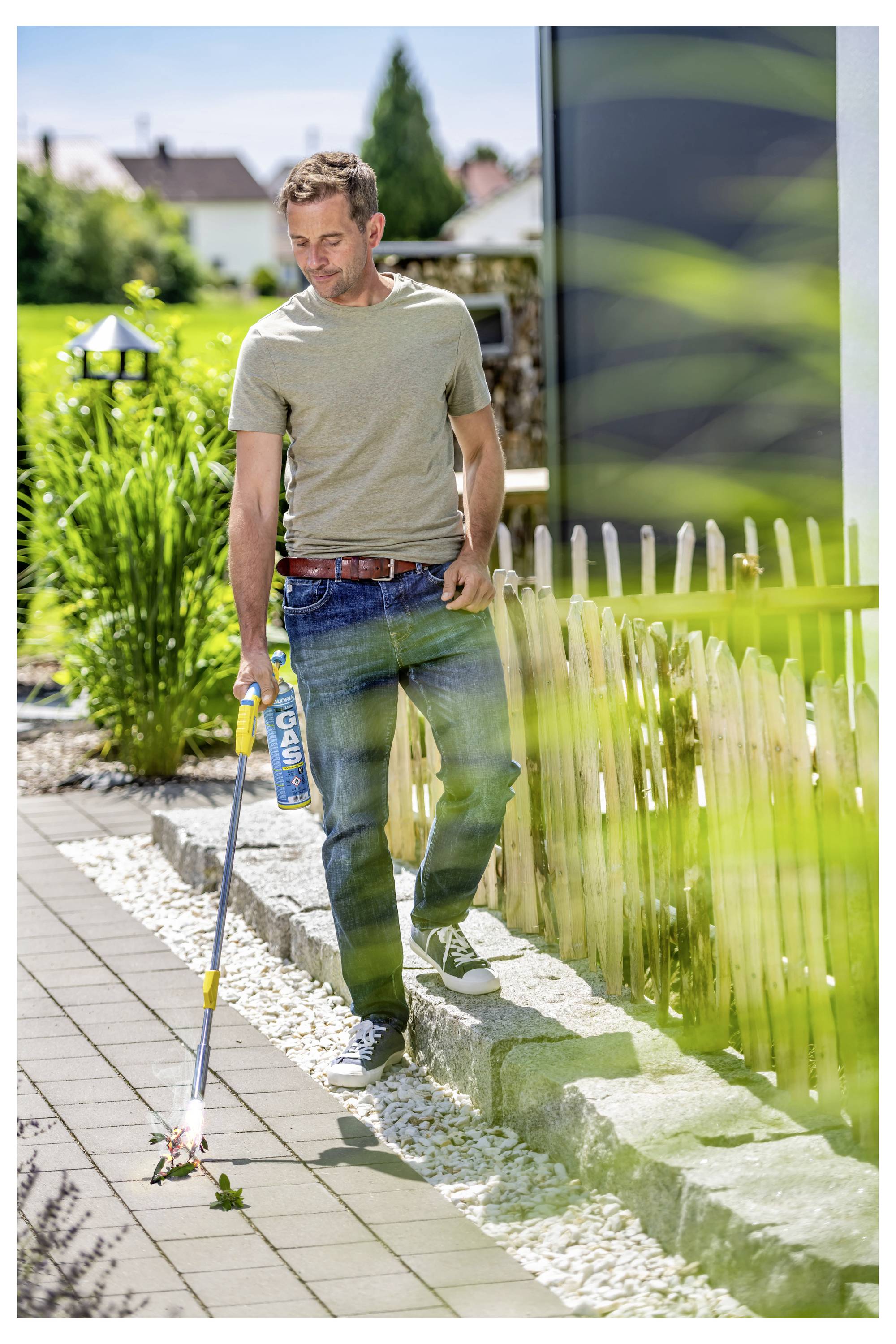 Ein Mann mit einem Unkrautvernichter beseitigt Unkraut vom Rand eines Gartenwegs neben einem Holzzaun an einem sonnigen Tag.
