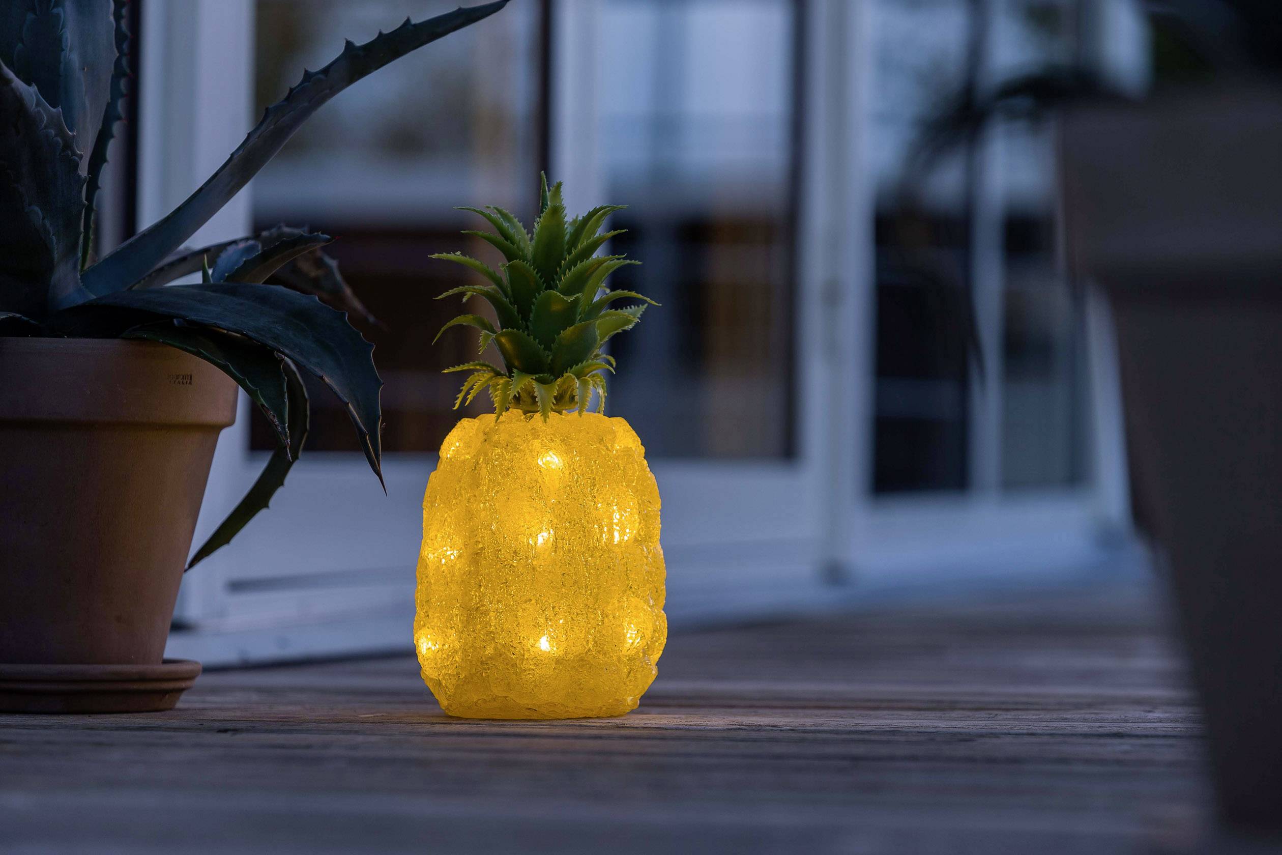 Eine leuchtende Lampe in Form einer Ananas steht auf einer Terrasse neben einer Pflanze, im Hintergrund ist eine Hausfassade bei Dämmerung.