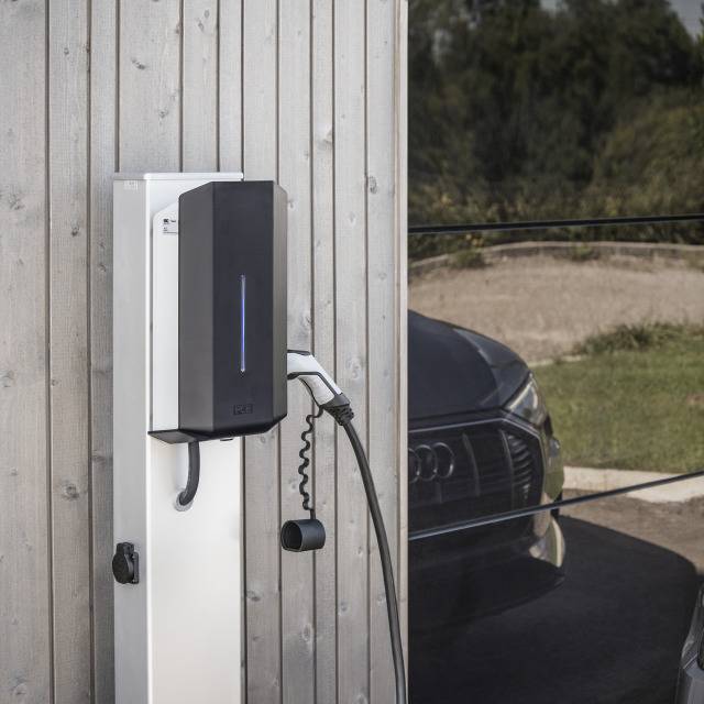 Elektroauto wird an einer Ladestation aufgeladen. Im Hintergrund ist ein Auto und eine Straße zu sehen.