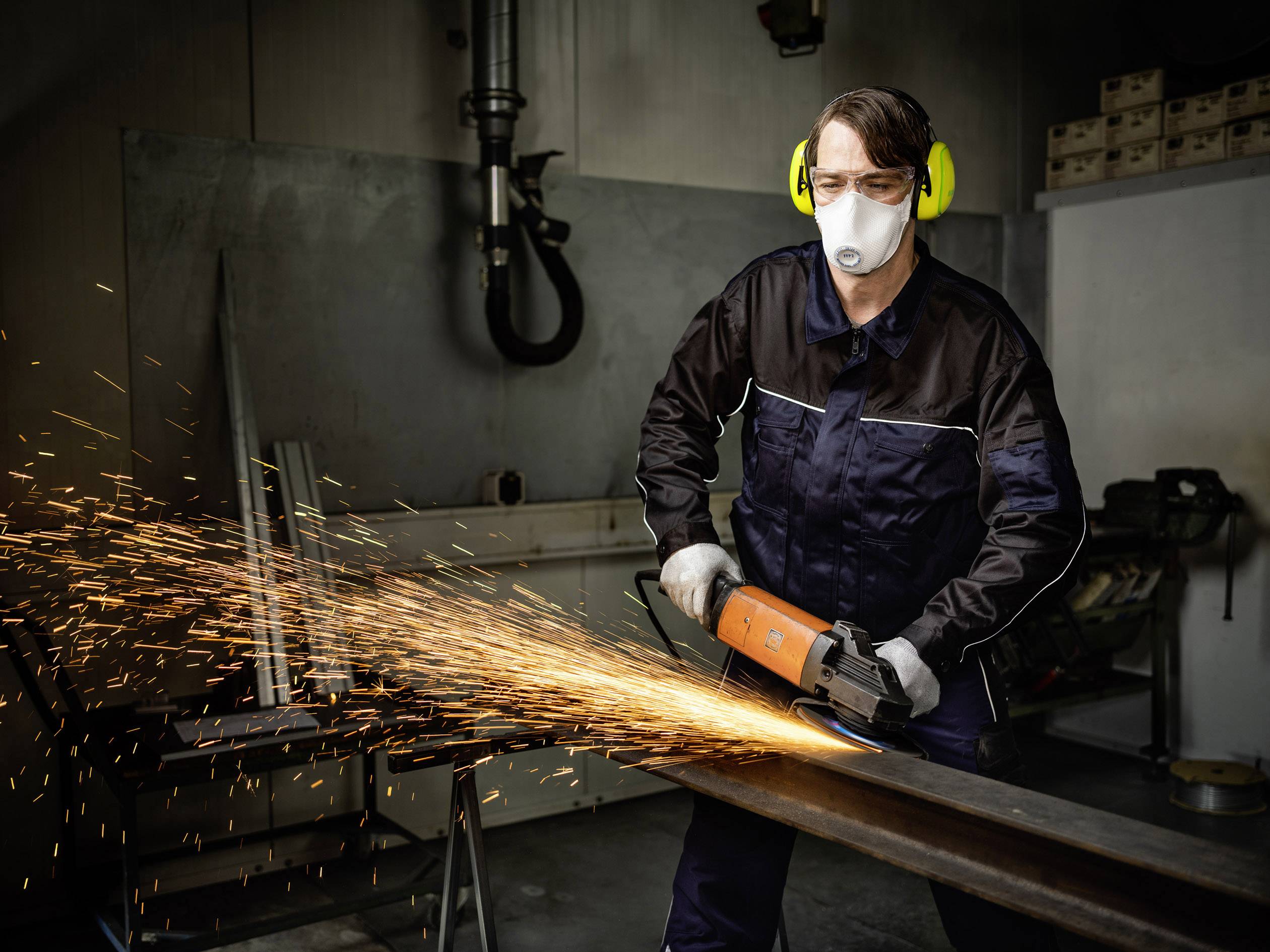 Ein Arbeiter mit Gehörschutz und Maske schleift Metall mit einem Winkelschleifer, Funken fliegen in einer Werkstatt.