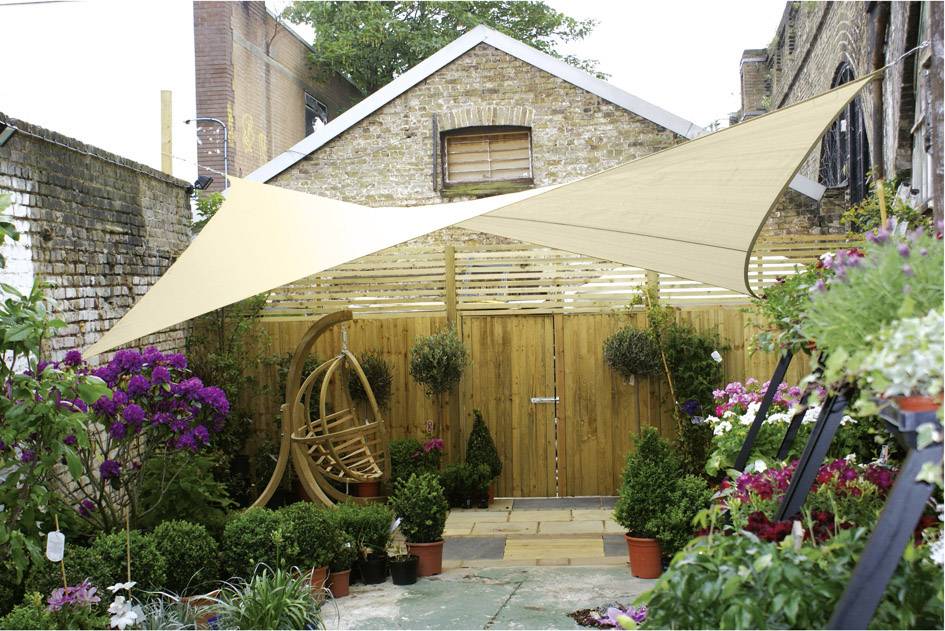 Ein sonniger Innenhof mit Holzschuppen, hölzernem Sichtschutz und Hängesessel, umgeben von blühenden Pflanzen und Sonnensegel.