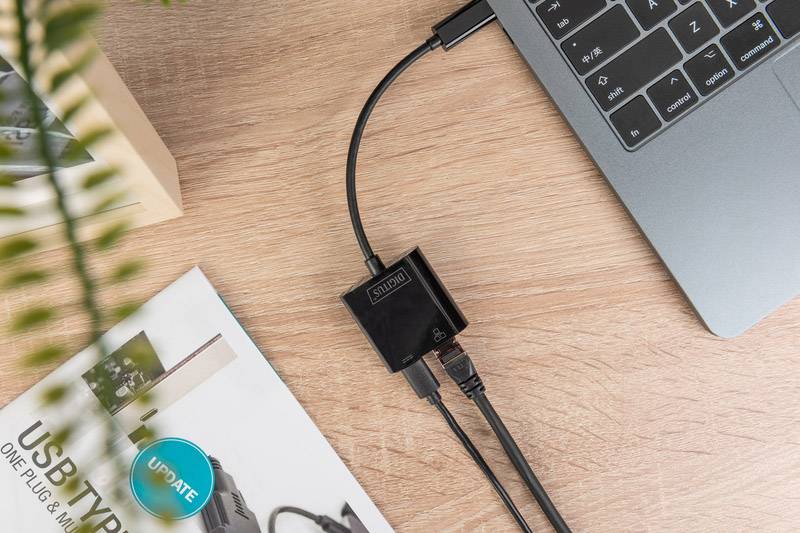 'Drei-in-eins-USB-C-Hub verbindet Laptop mit mehreren Geräten auf Holztisch. Neben dem Hub liegt eine Zeitschrift und eine Pflanze.'