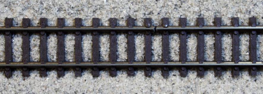 Eine Nahaufnahme eines Modellbahngleises auf einem kiesartigen Untergrund, das die Details der Schienen und Schwellen zeigt.