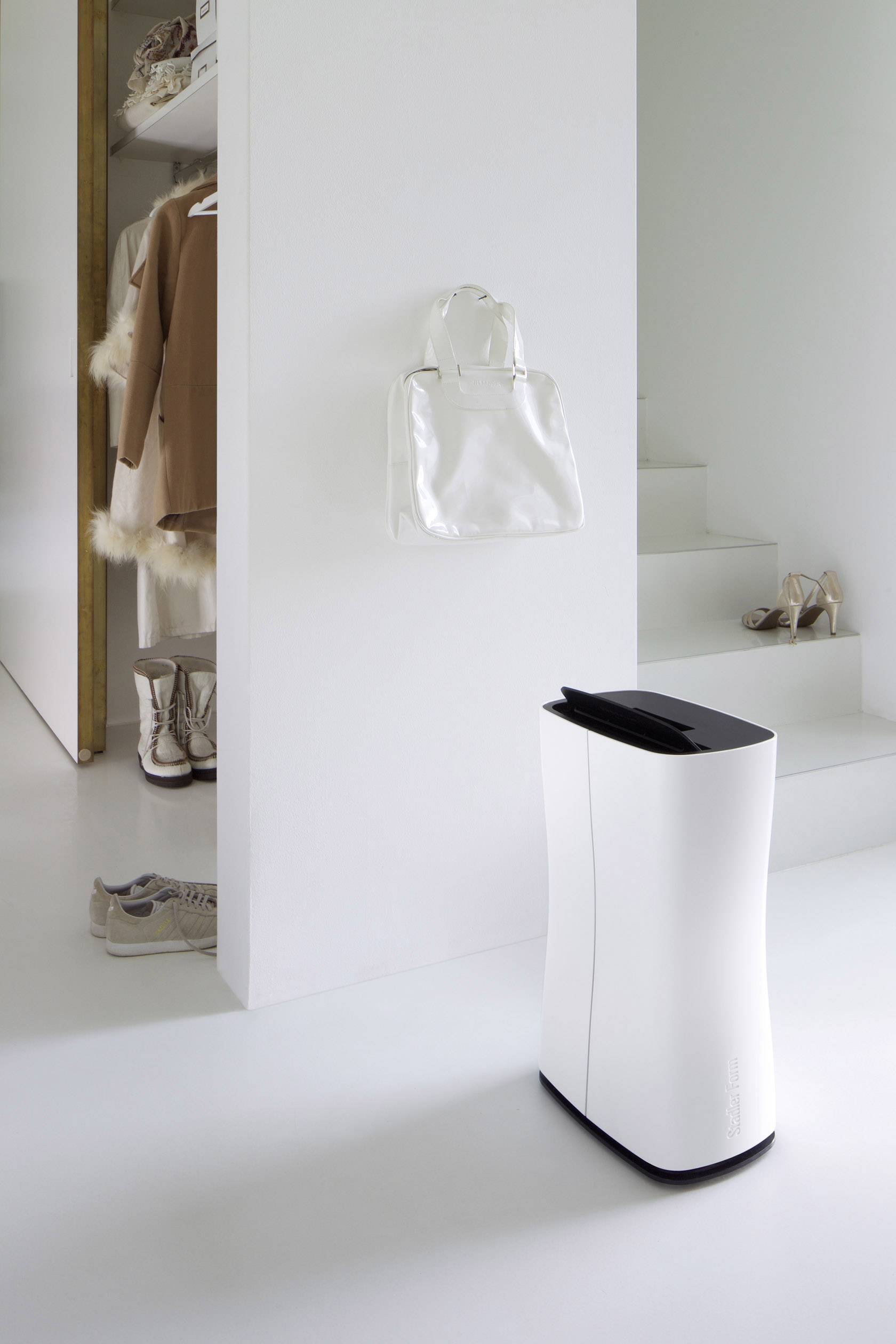Ein weißer Luftreiniger steht vor einer weißen Wand in einem modernen, minimalistischen Raum mit Schuhen und einer Handtasche.