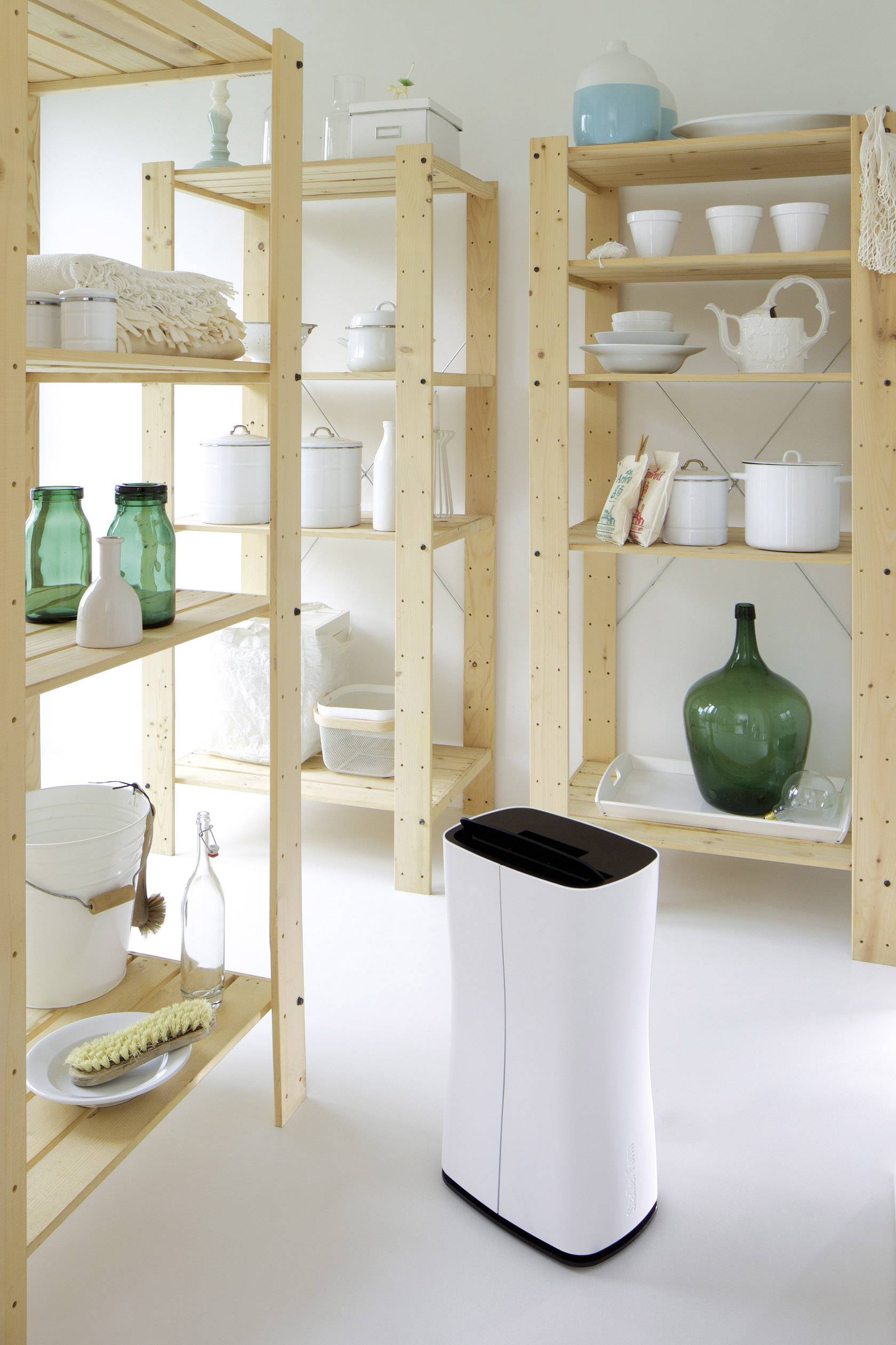 Ein weißer Luftentfeuchter steht in einem Holzregal-Zimmer mit Keramik, Glasflaschen und Haushaltsgegenständen.