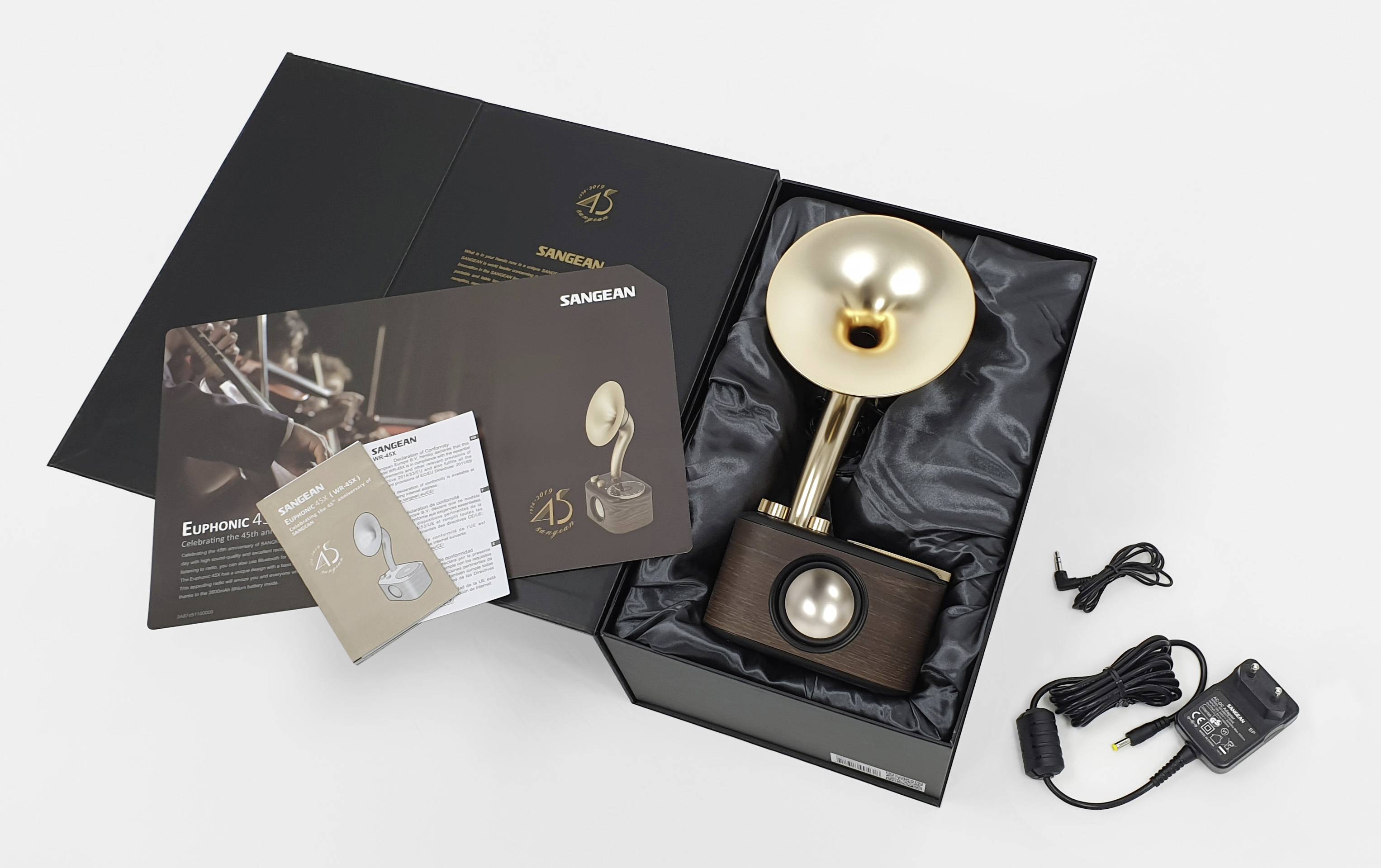 Eine geöffnete Geschenkbox enthält einen goldenen Retro-Lautsprecher in Grammophon-Form, mit beigelegten Broschüren und einem Ladegerät.
