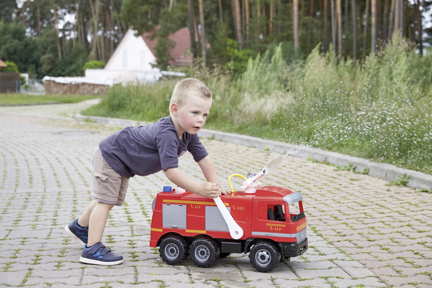 Ein kleiner Junge spielt konzentriert auf einer gepflasterten Fläche mit einem Spielzeug-Feuerwehrauto. Im Hintergrund sind Bäume zu sehen.
