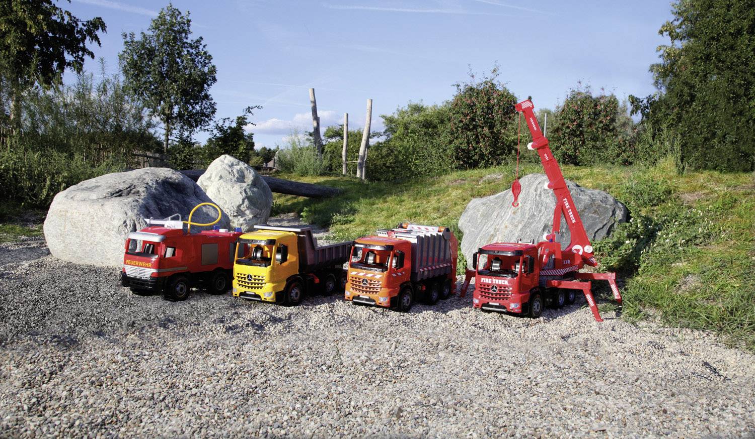 Vier bunte Spielzeuglastwagen stehen nebeneinander auf einem Kiesweg im Freien, umgeben von Bäumen und großen Steinen.