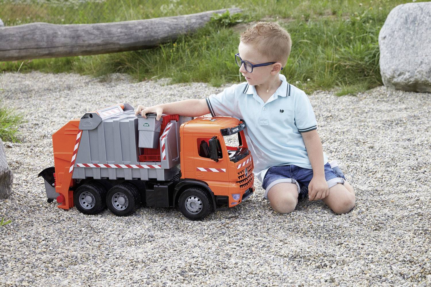 Ein Junge in blauer Kleidung spielt im Freien mit einem großen Spielzeug-Müllwagen auf einem Kiesweg.