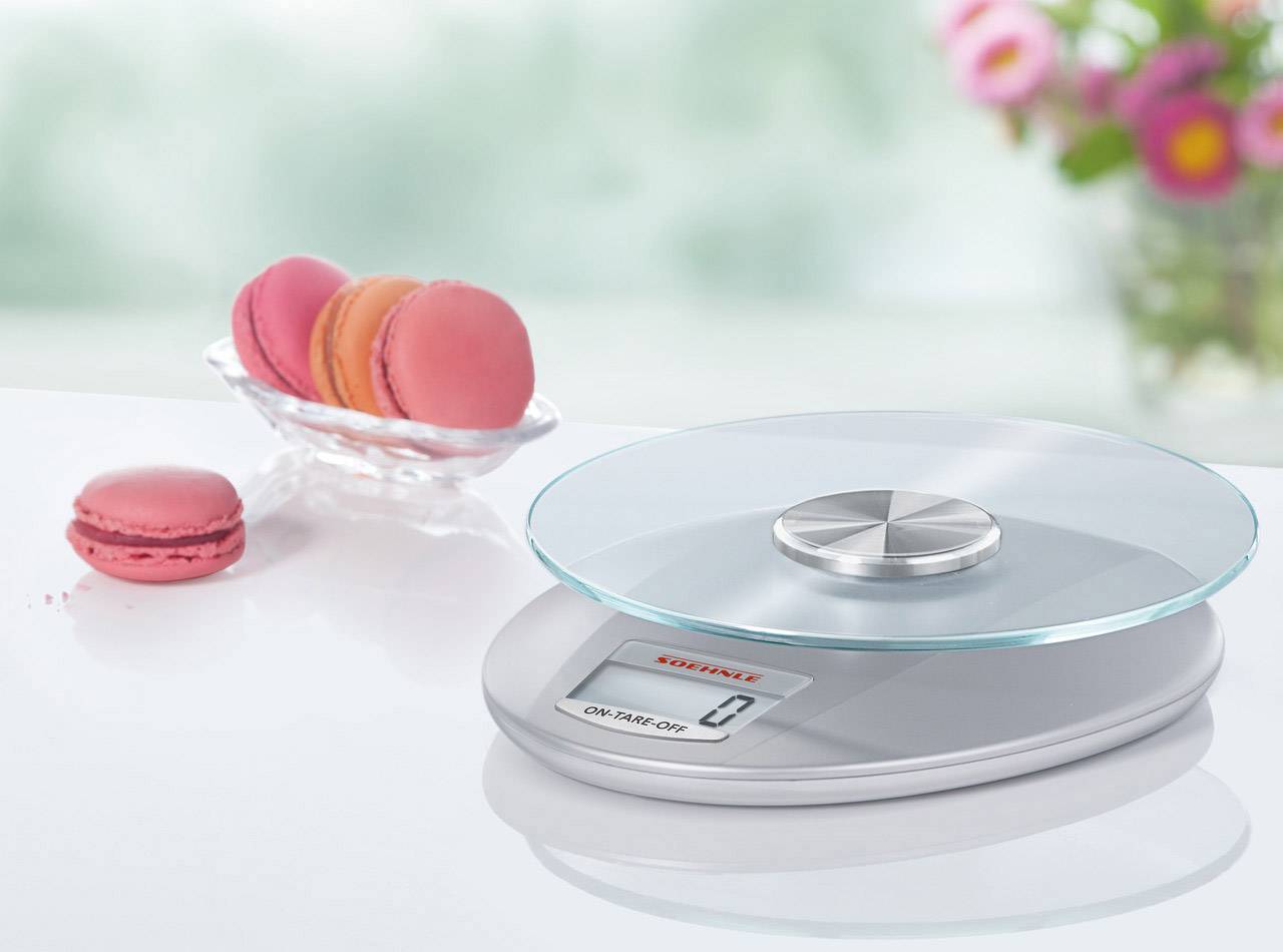 Elektronische Küchenwaage auf einem Tisch, zeigt '0 g'. Daneben eine Schale mit rosa Macarons. Im Hintergrund verschwommene Pflanzen.