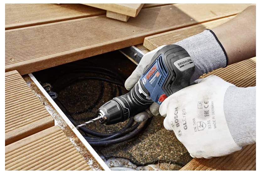 Eine Person verwendet eine kabellose Elektrobohrmaschine, um Holzterrassendielen zu befestigen. Die Bohrmaschine ist blau und schwarz, und die Person trägt Schutzhandschuhe.