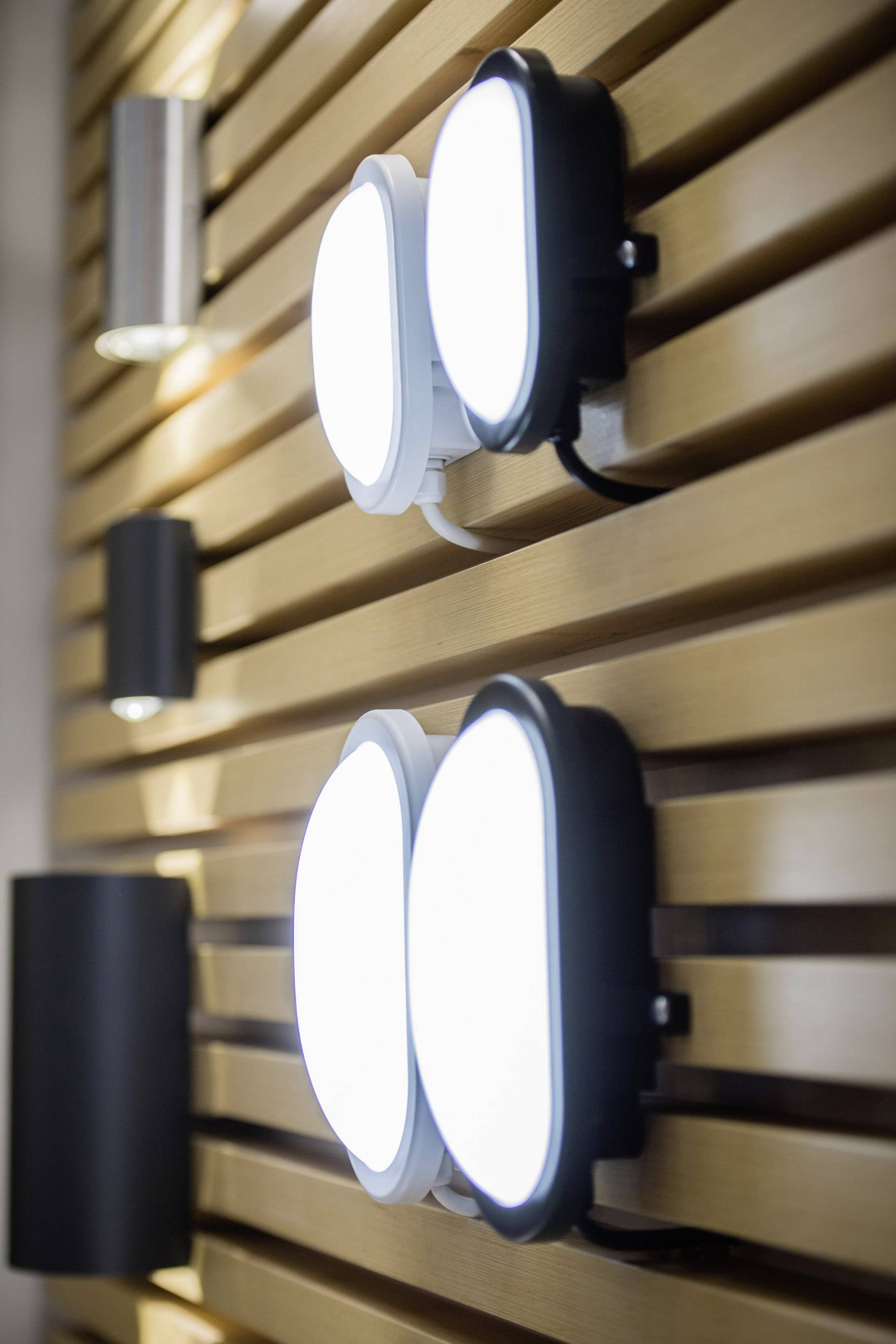 Weiße und schwarze ovale Leuchten sind symmetrisch an einer Wand aus Holzlamellen montiert und strahlen helles Licht aus.