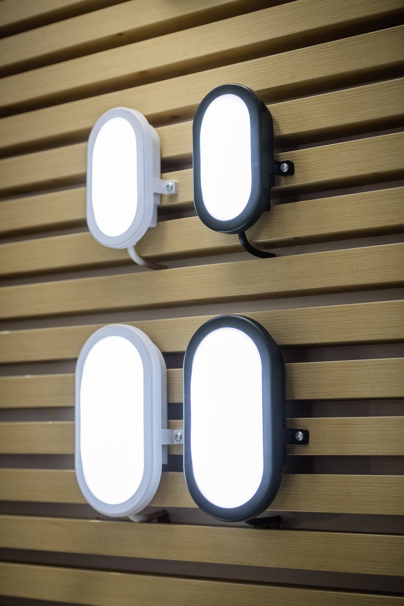 Vier ovale Wandlampen, je zwei weiße und zwei schwarze, sind an einer Holzlamellenwand befestigt. Lampen leuchten hell.