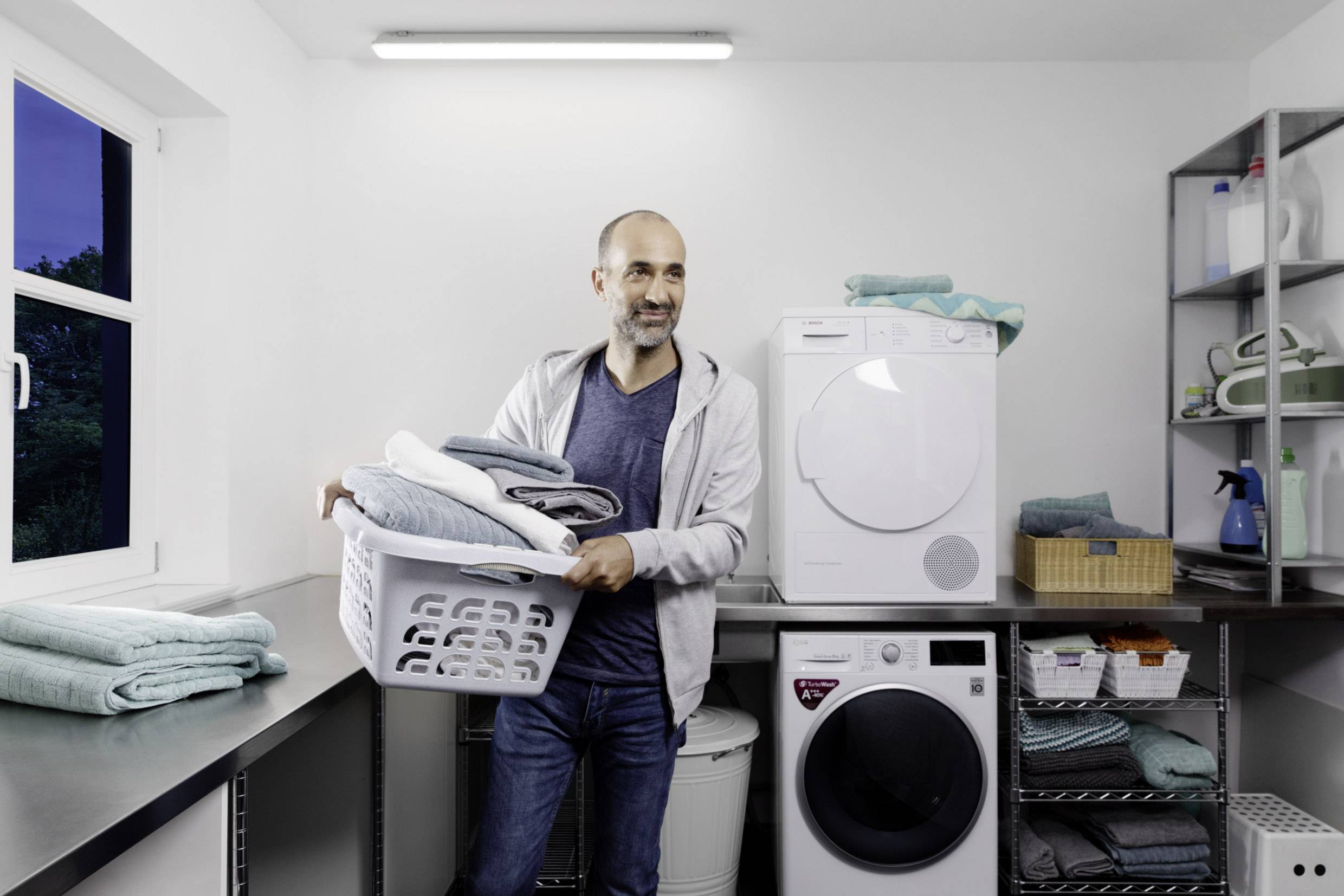 Ein Mann trägt einen Wäschekorb voller zusammengelegter Kleidung in einer Waschküche mit Waschmaschine, Trockner und Regalen.
