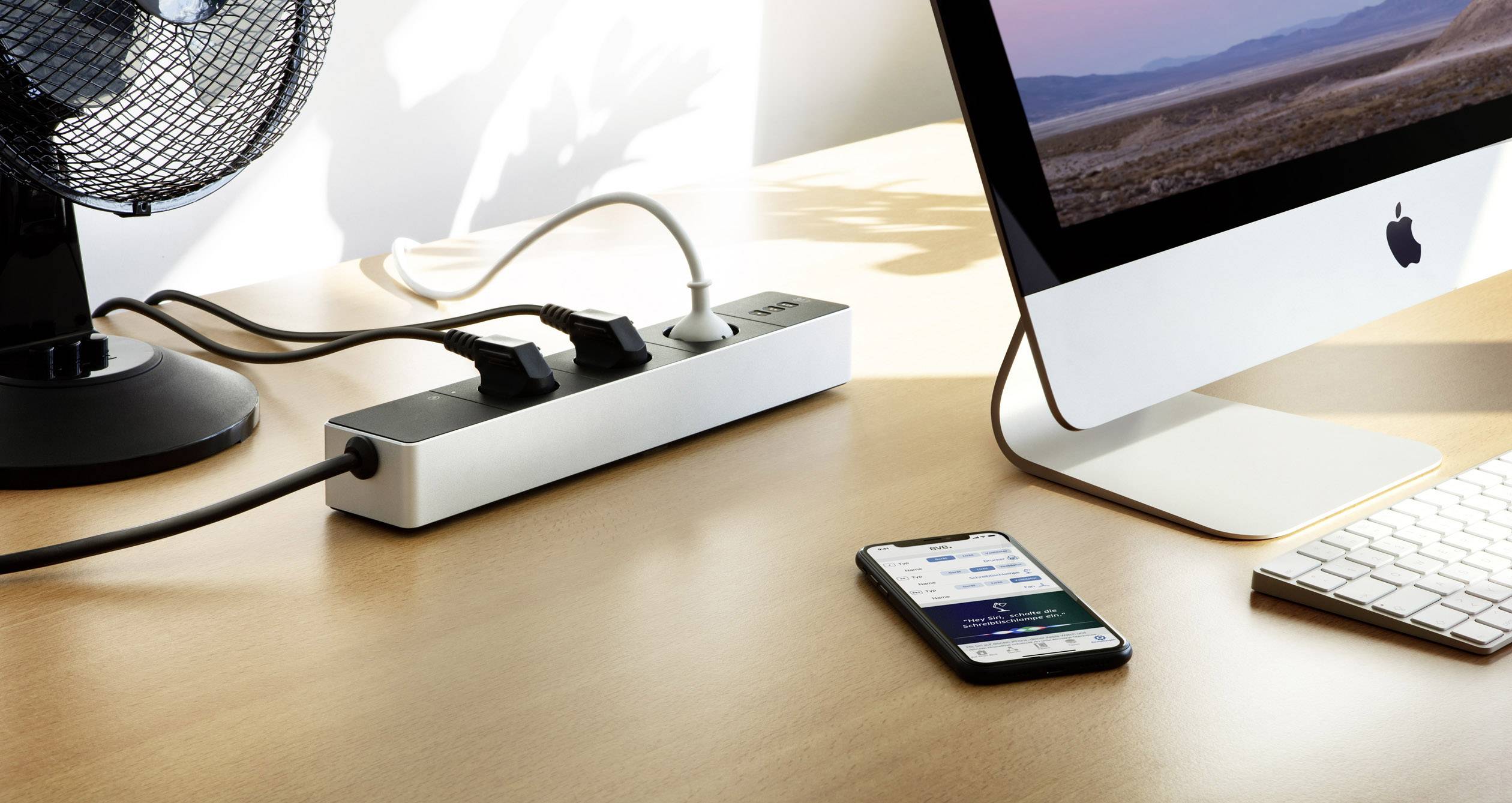 Ein Schreibtisch mit einem iMac, einer Mehrfachsteckdose und einem Smartphone. Die Steckdose ist mit mehreren Geräten verbunden.