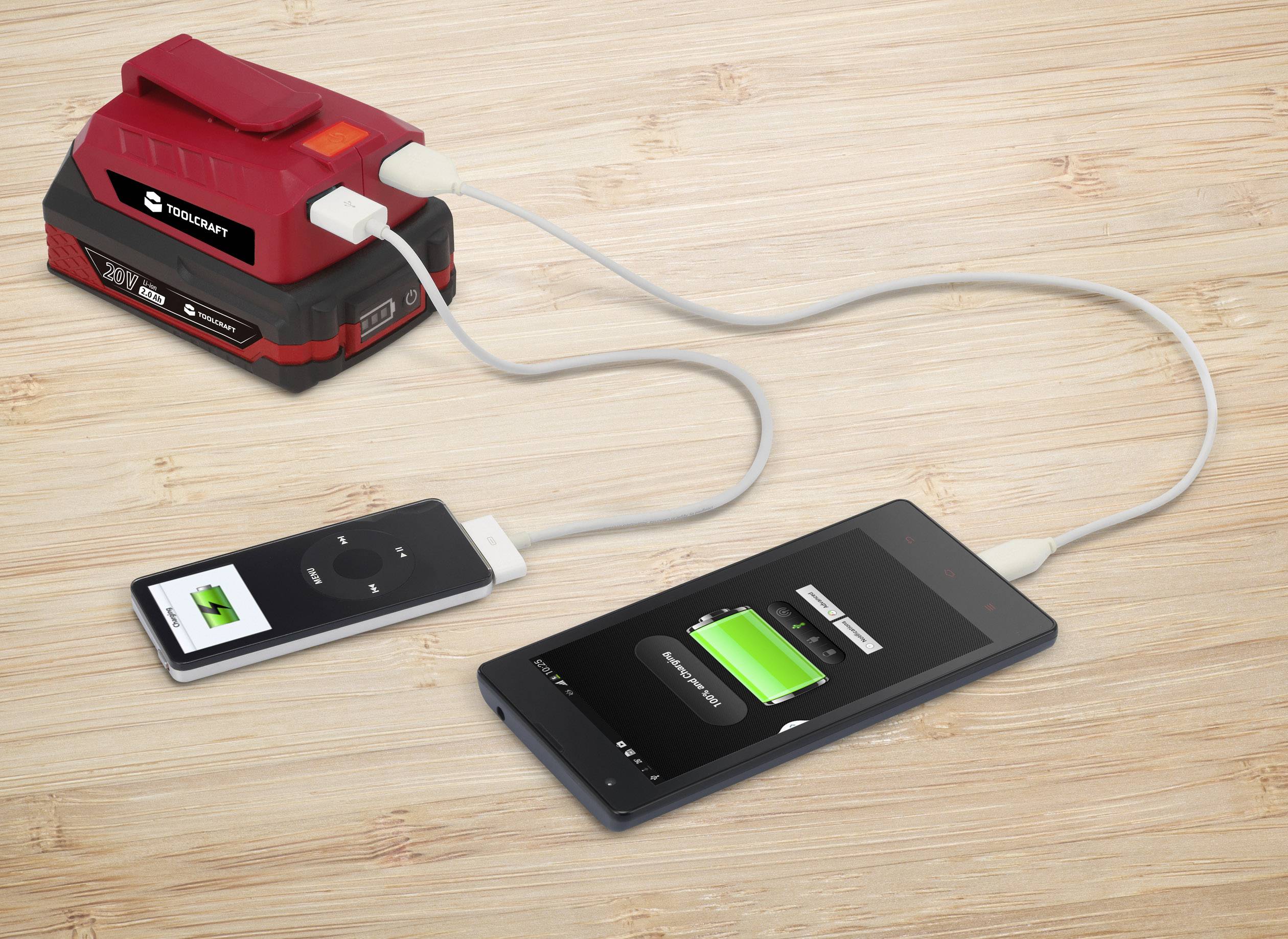 Auf einem Holztisch lädt ein roter Akkupack zwei elektronische Geräte über USB-Kabel. Beide Geräte zeigen Ladeanzeigen.