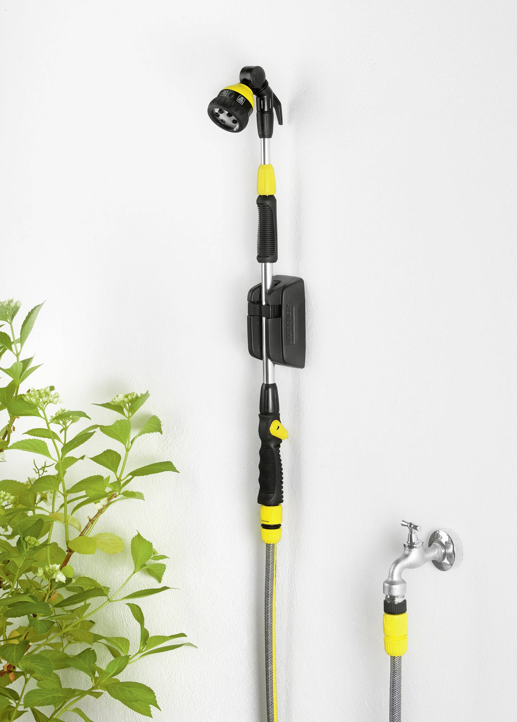 Duschkopf mit gelbem und schwarzem Griff an einer Wand befestigt. Rechts daneben ein Wasserhahn und links ein grüner Pflanzenzweig.