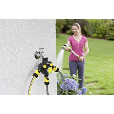Eine Frau gießt den Garten mit einem Schlauch. Im Vordergrund ist ein an der Wand montierter Wasserverteiler mit mehreren Anschlüssen zu sehen.