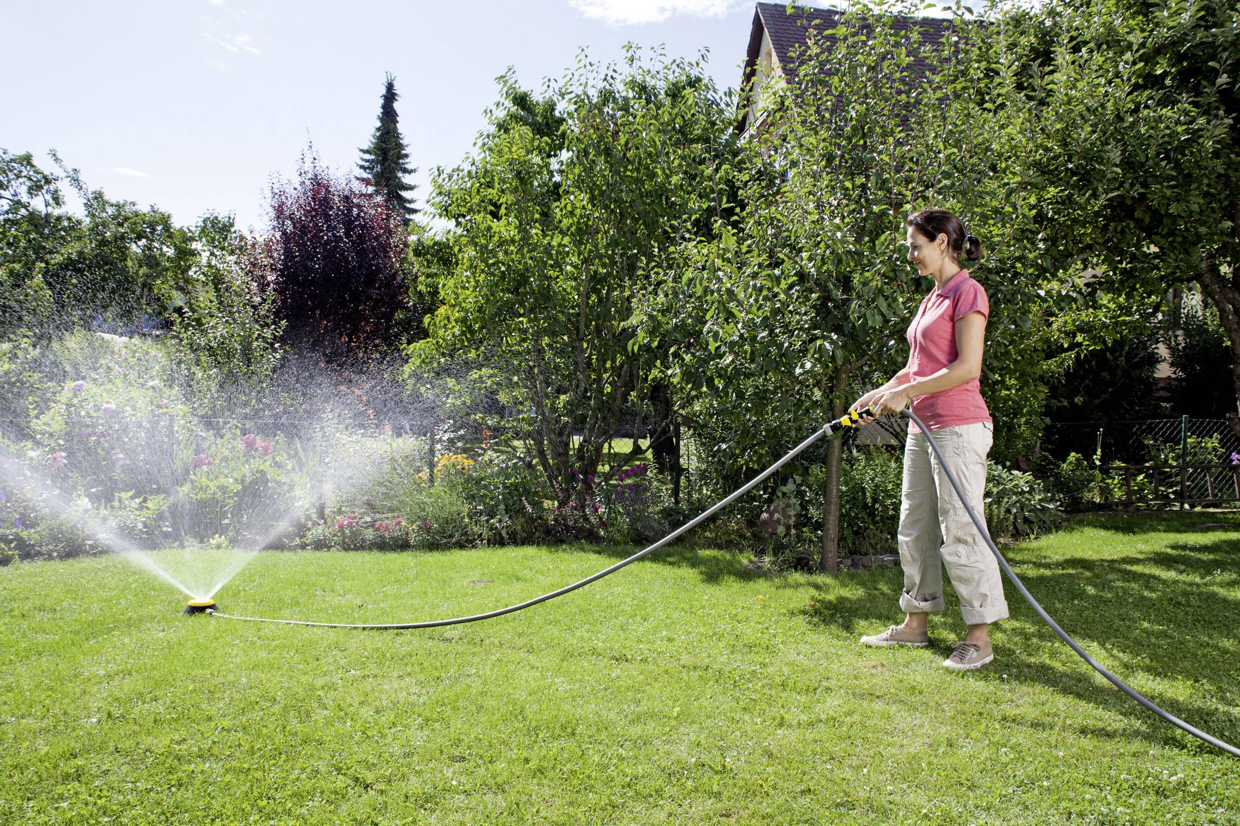 Eine Frau bewässert mit einem Schlauch einen grünen Rasen im Garten. Im Hintergrund sind Bäume und ein blauer Himmel zu sehen.